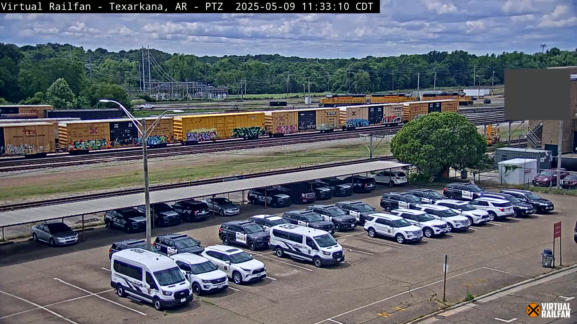 A mostly sunny day shows a long line of graffiti-covered train cars parked next to a lot full of police vehicles under a covered parking structure.