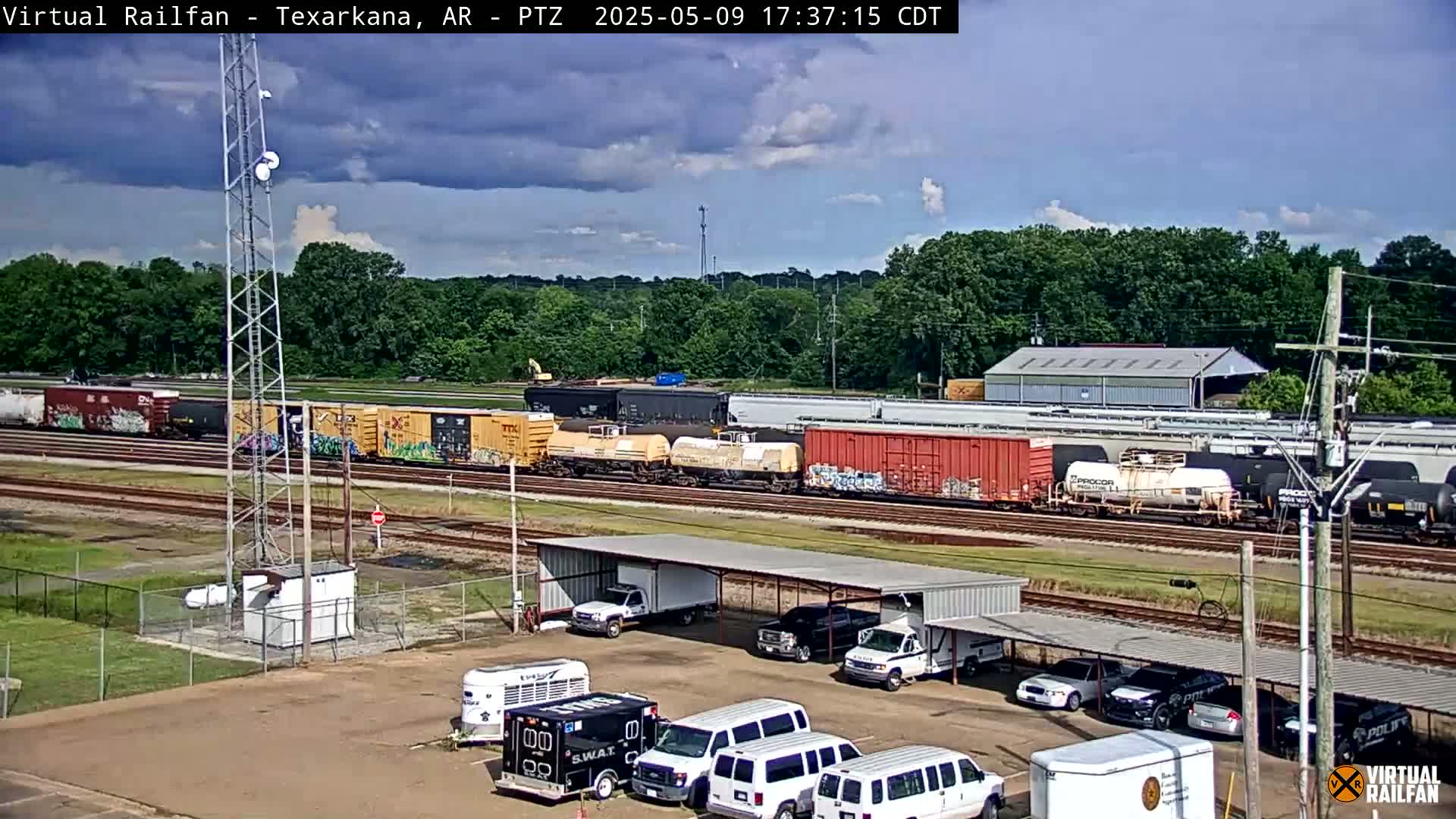A long freight train sits on tracks near a parking lot with several vehicles, under a partly cloudy sky.