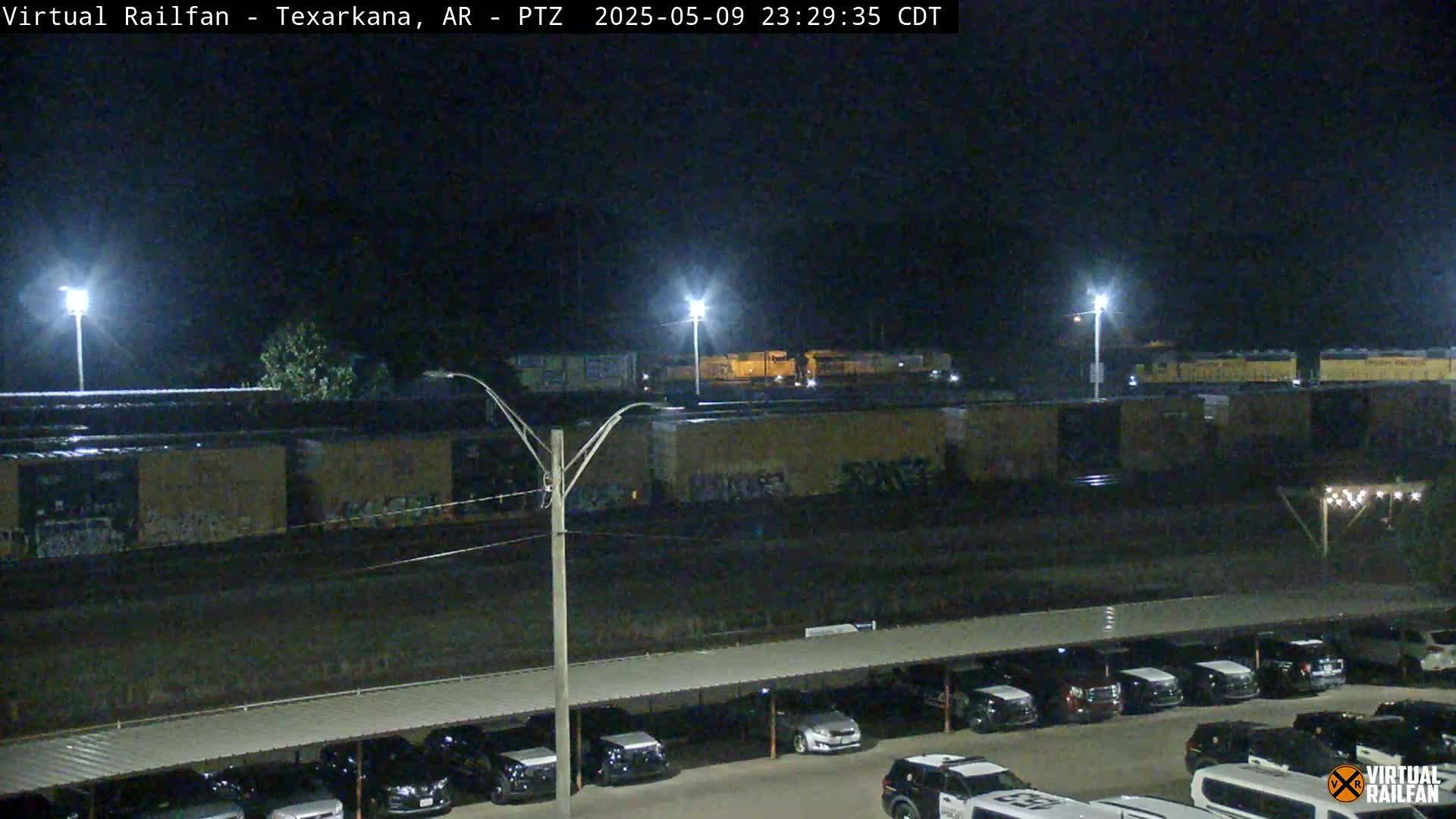 A nighttime view of a train yard with several boxcars, a parking lot with cars under a covered structure, and bright lights illuminating the scene.