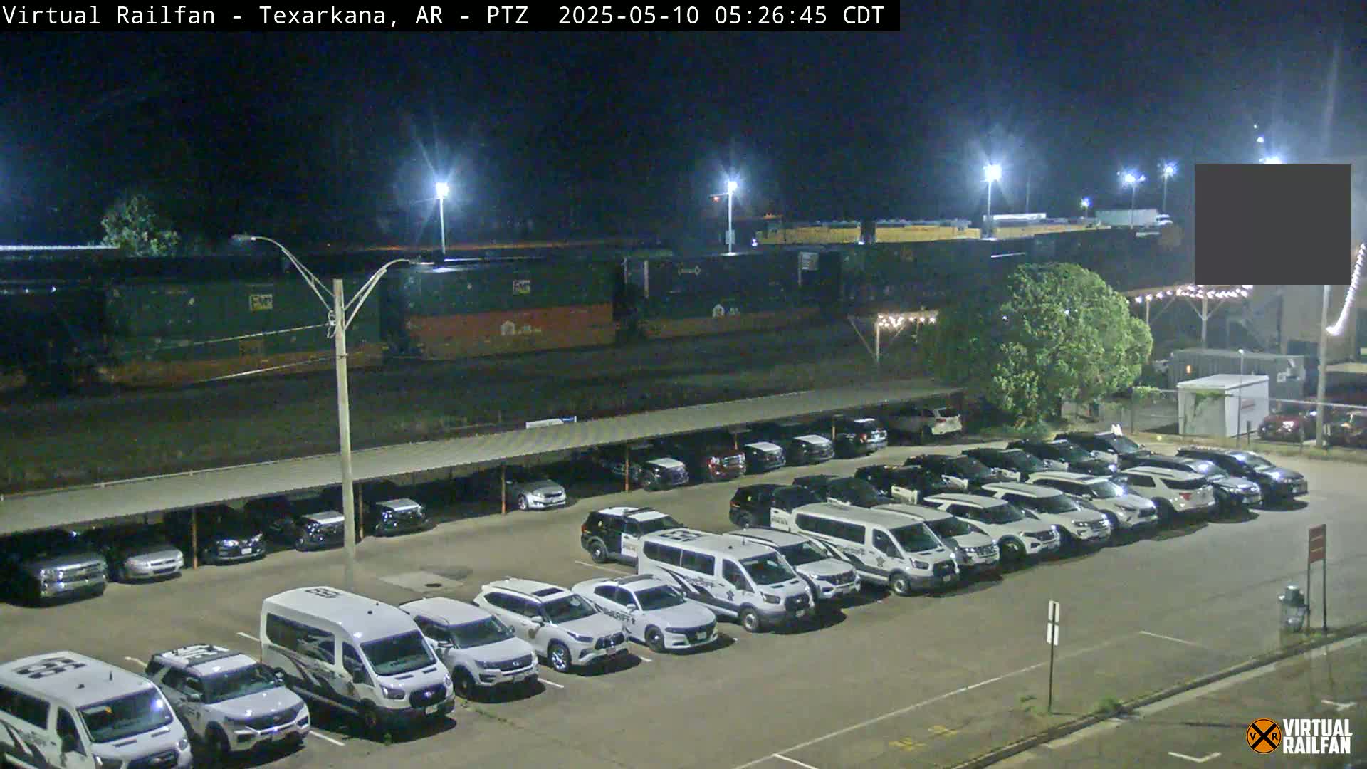 At night, a long freight train sits in a rail yard adjacent to a parking lot full of cars and vans, some of which appear to be law enforcement vehicles.