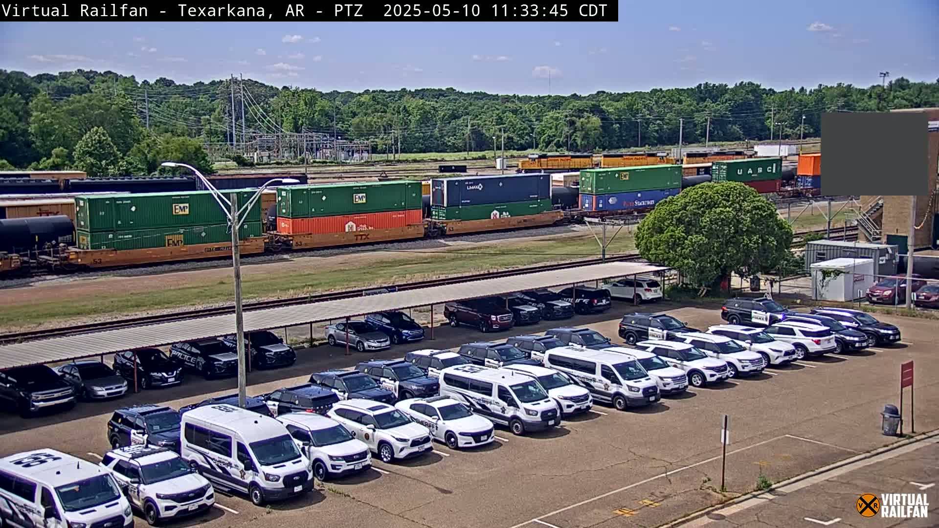 Under a sunny sky, a freight train carrying various shipping containers sits in a rail yard next to a parking lot full of police vehicles parked under a covered structure.