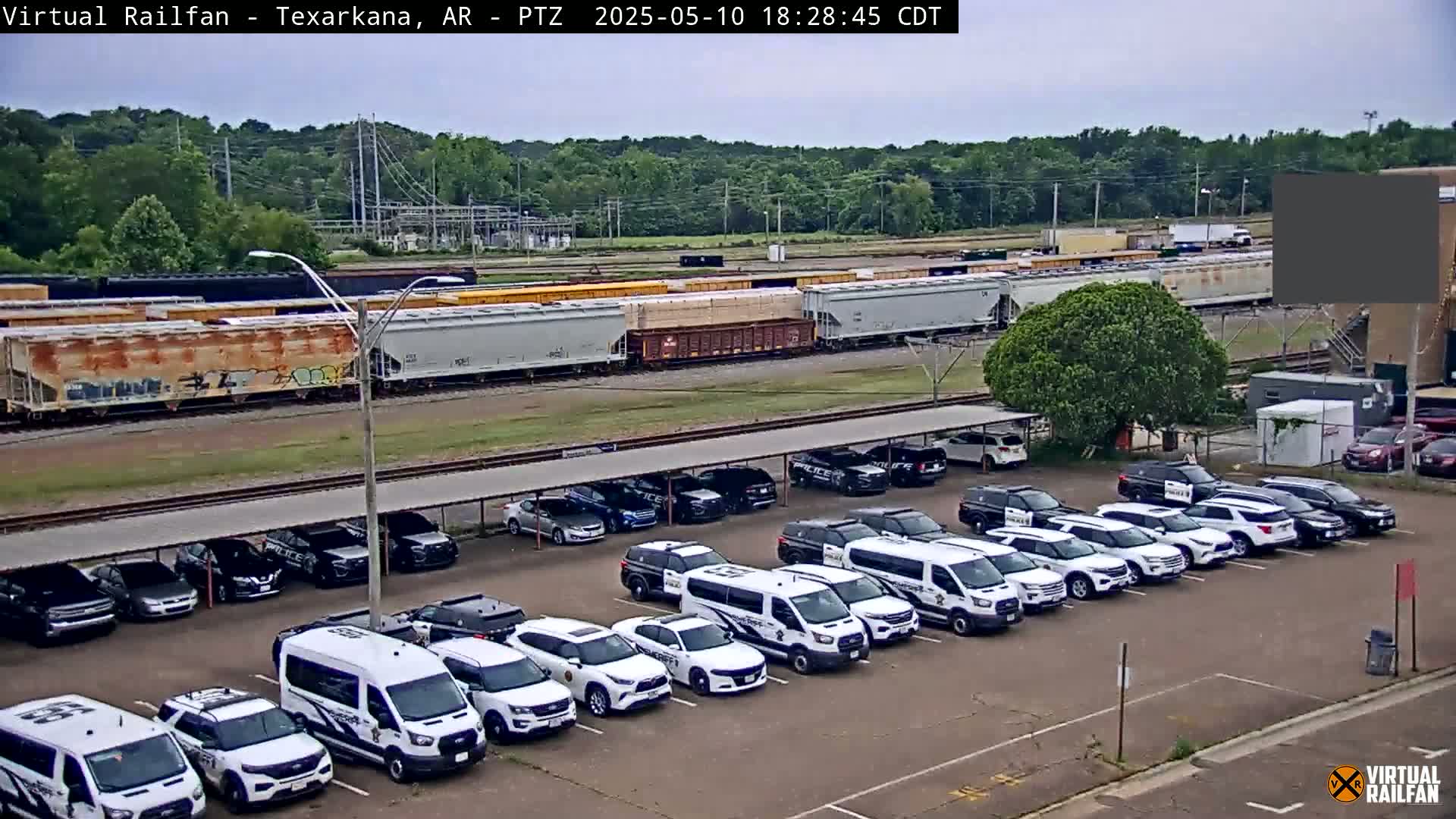 A long freight train is parked near a lot filled with mostly white police vehicles under a covered parking structure, on an overcast day.