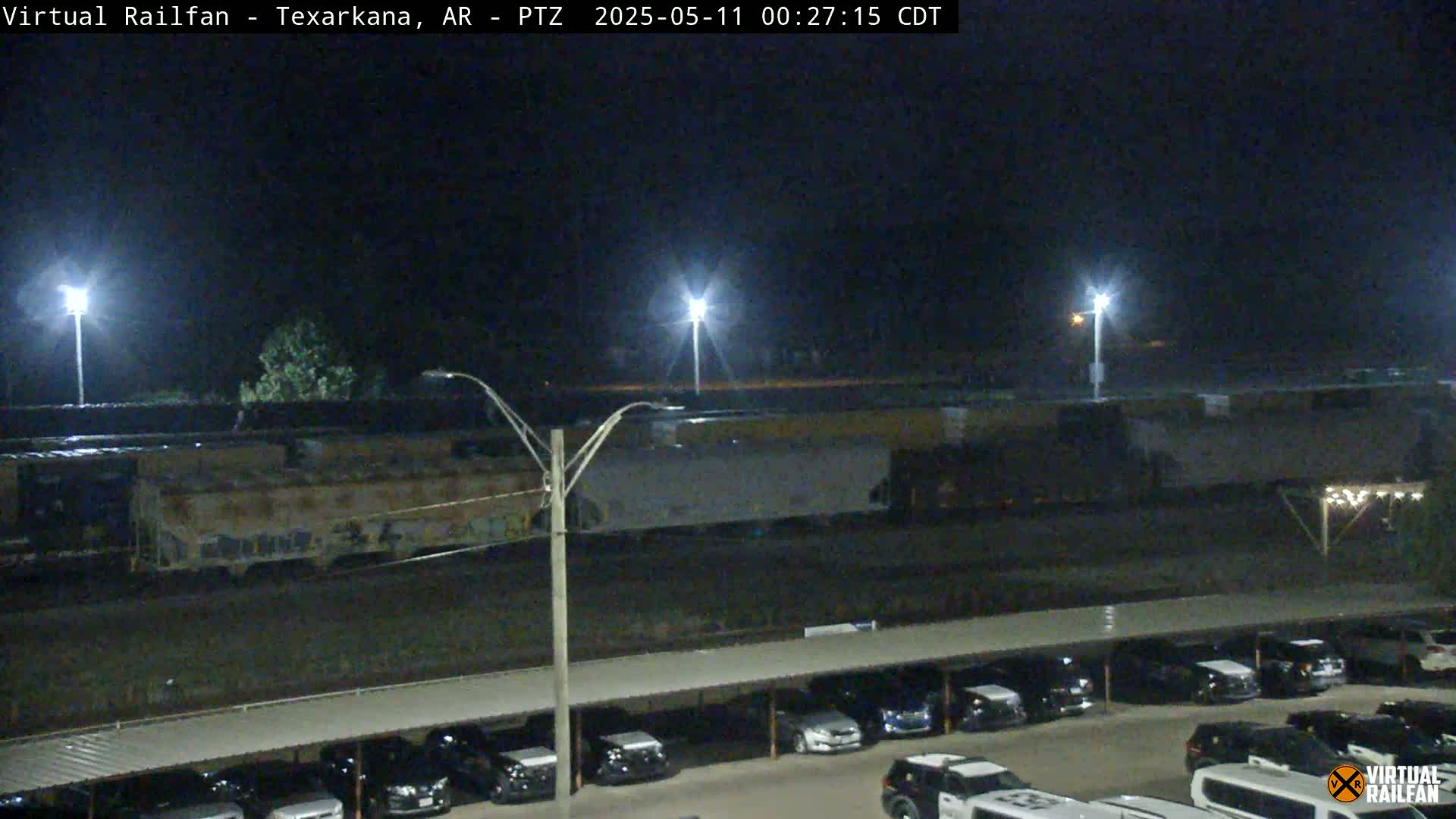 At night, a long freight train sits near a parking lot under a covered structure, illuminated by several bright lights.