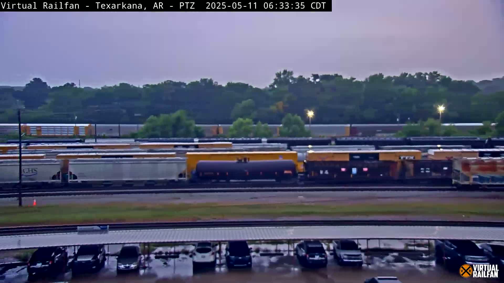A long train of freight cars sits in a rail yard under an overcast sky, with a parking lot visible in the foreground and rain-slicked surfaces.