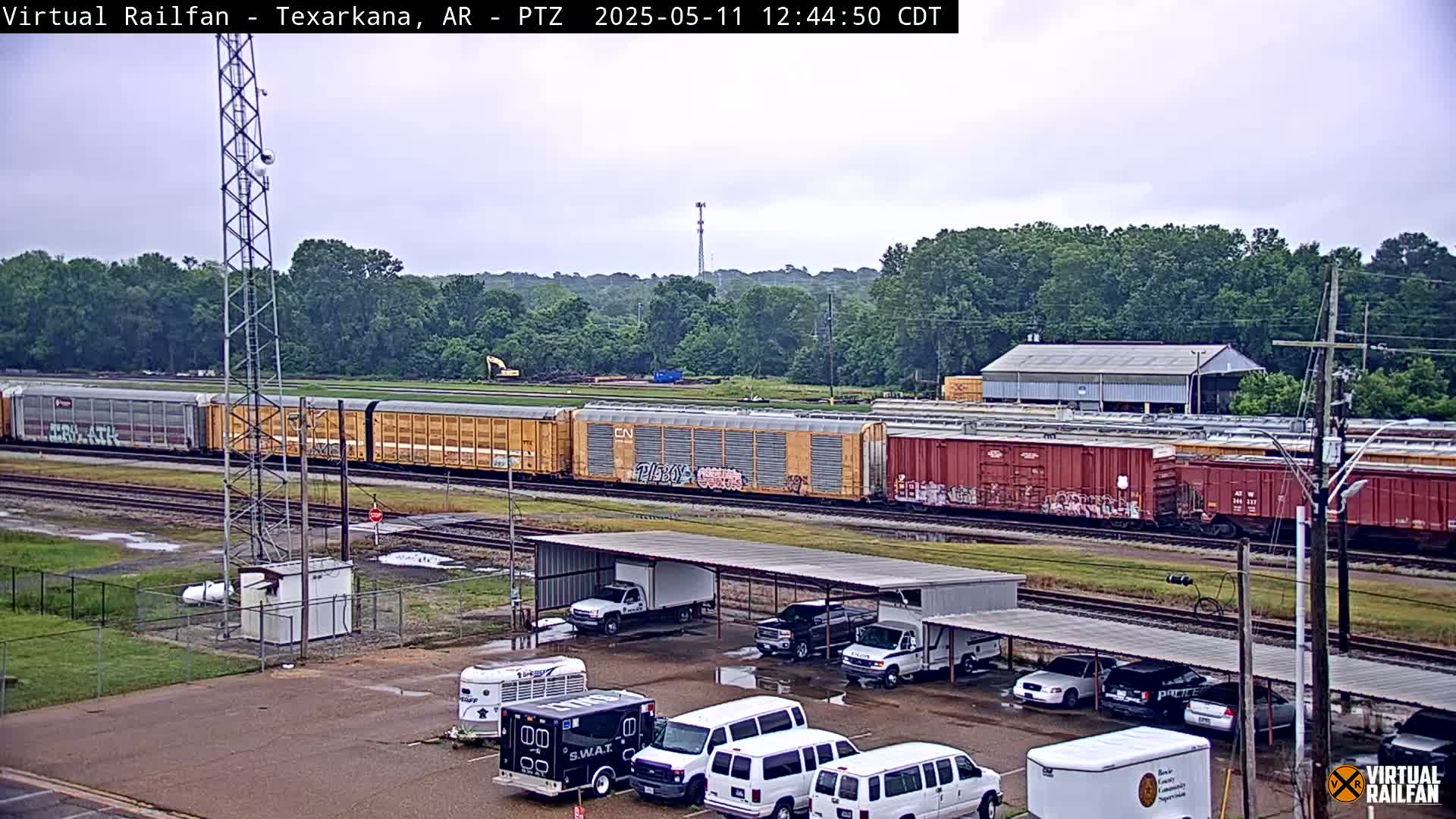 A long freight train passes by a parking lot with several vehicles under a covered structure, all situated beneath an overcast sky.
