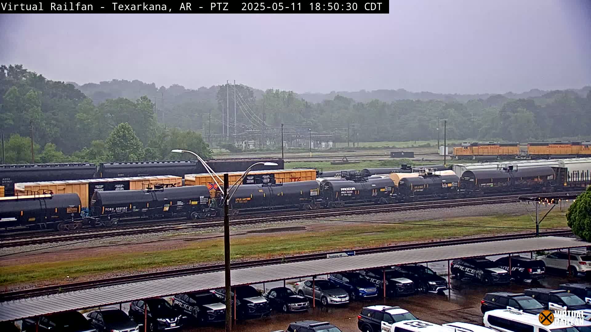 A long freight train sits on railroad tracks in the rain, parked next to a lot of police cars under a covered structure.