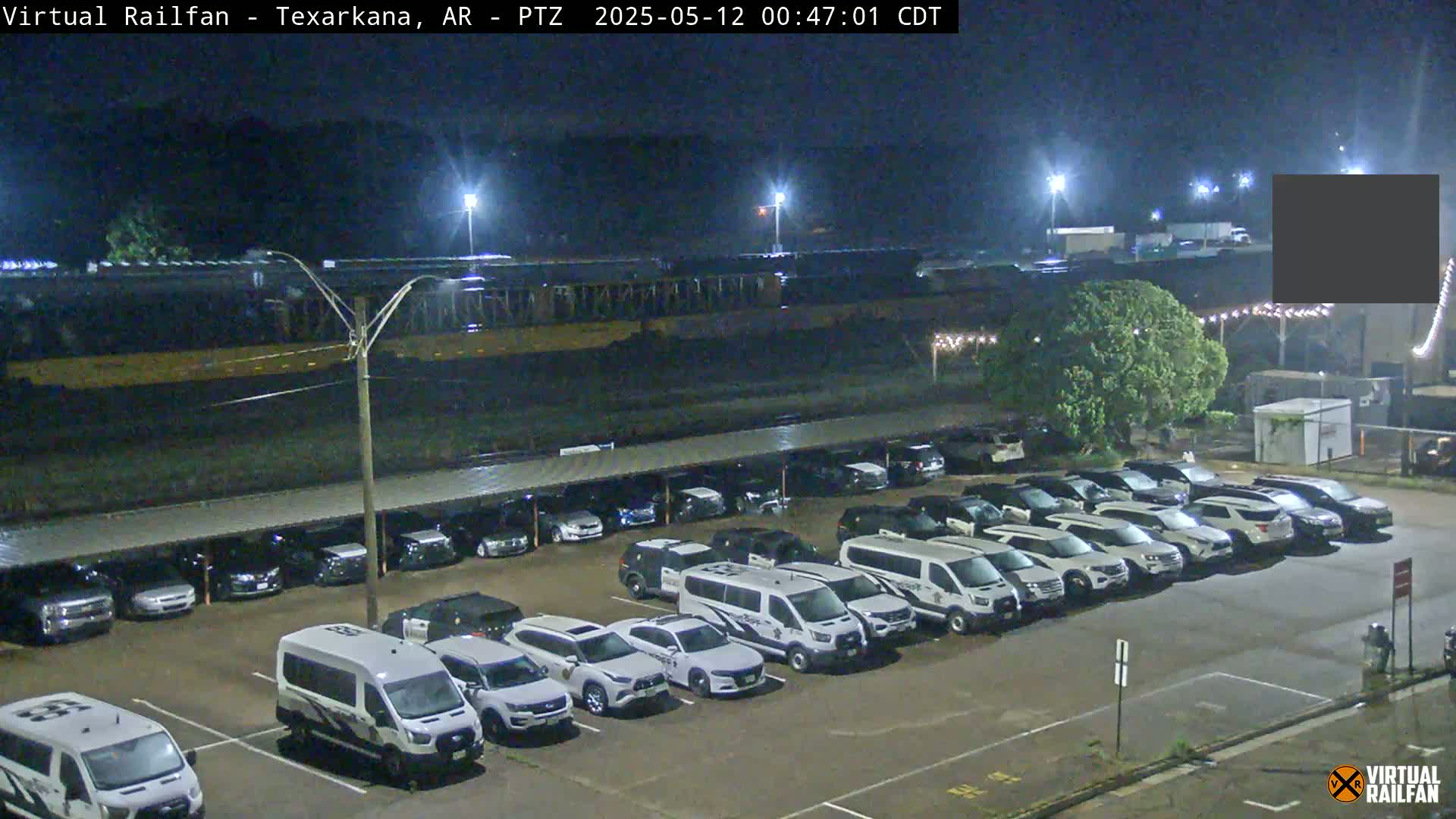 At night, a long freight train is parked next to a large parking lot filled with mostly white cars and vans under a covered structure.