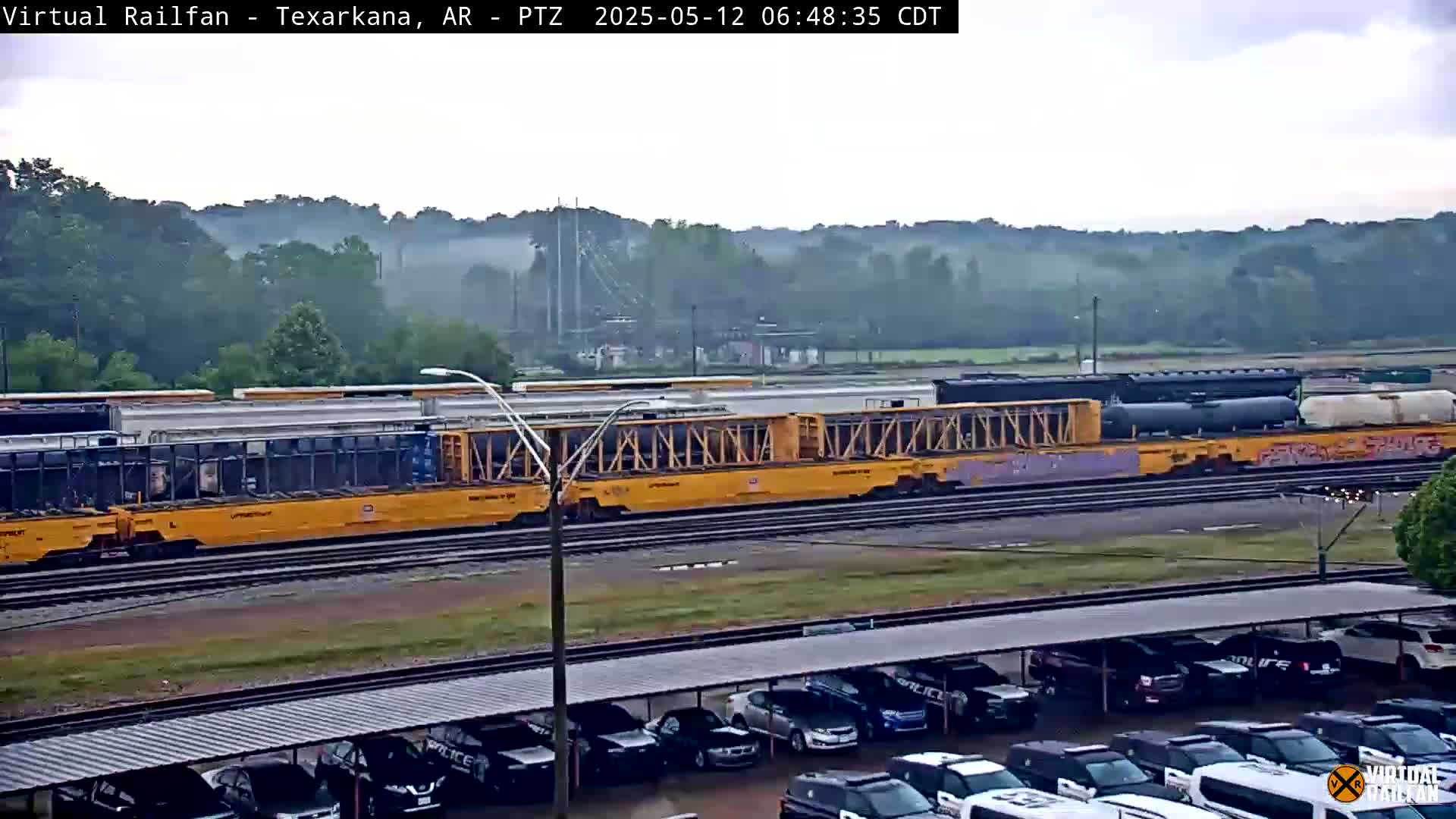 A long freight train sits on tracks in front of a parking lot under a misty sky.
