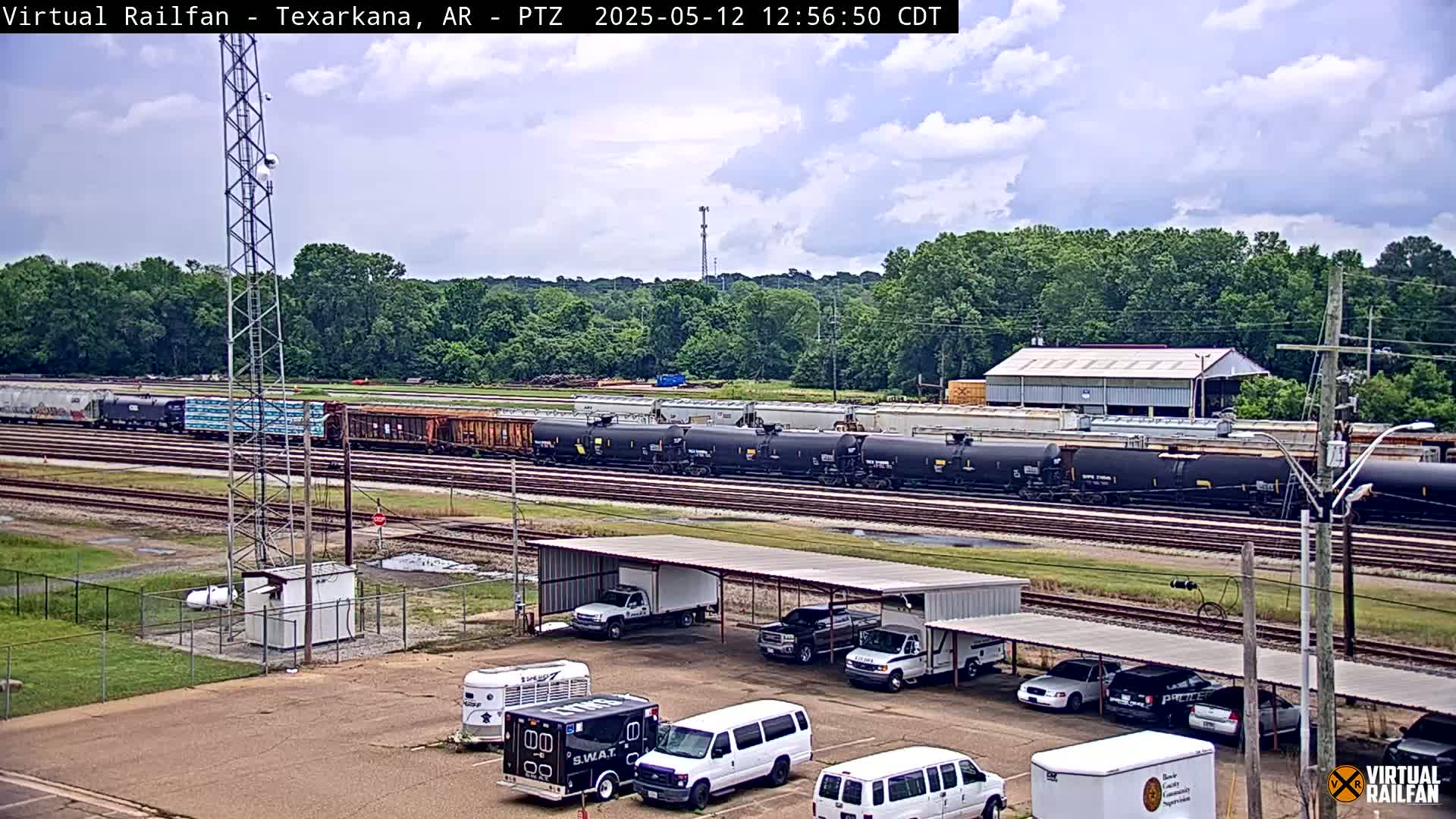 A long freight train sits on railroad tracks next to a parking lot with several vehicles under a covered structure, all under a partly cloudy sky.