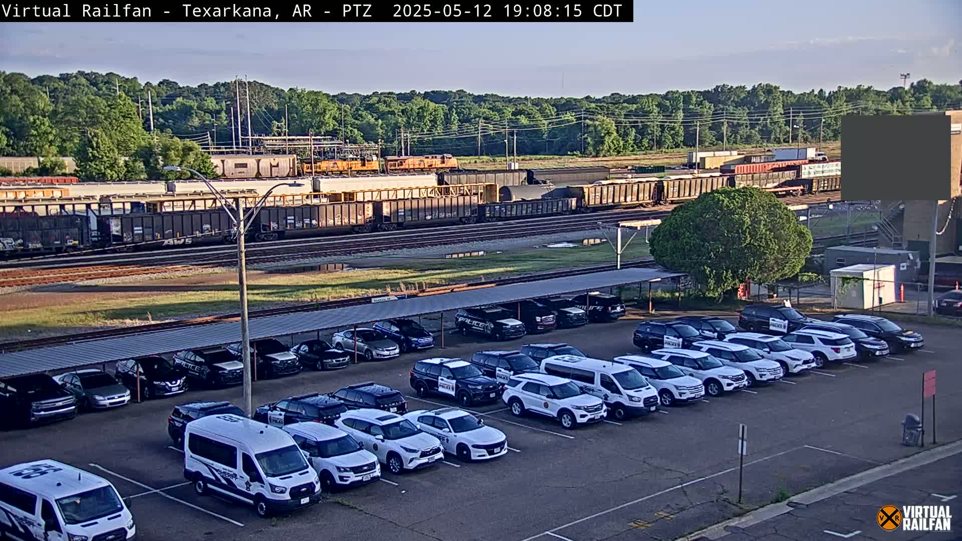 A long freight train sits on tracks behind a parking lot full of police vehicles under a covered structure, all under a partly cloudy sky.