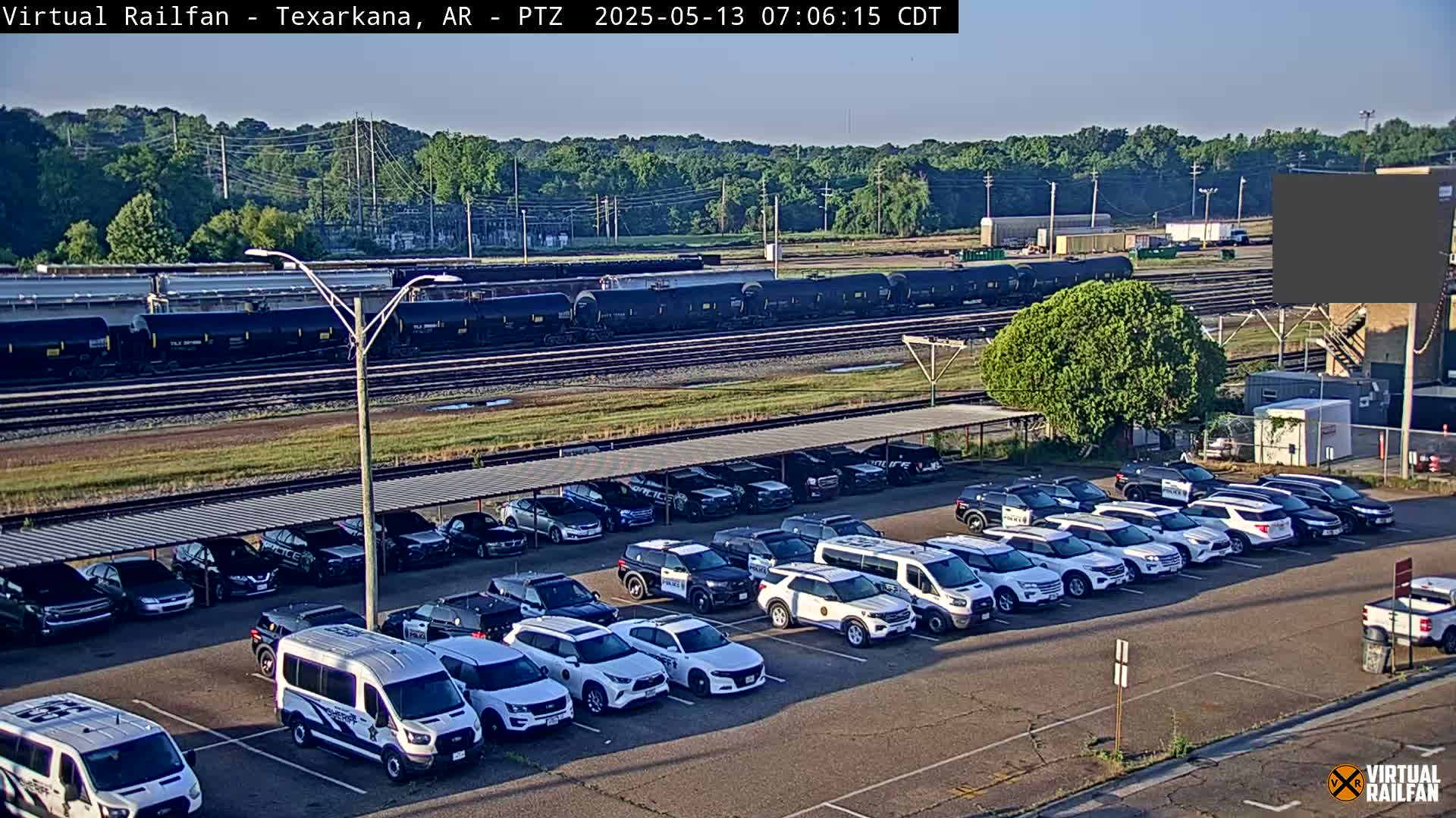 A long line of black train cars sits on tracks behind a large parking lot filled with police vehicles under a covered structure, all on a clear, sunny day.