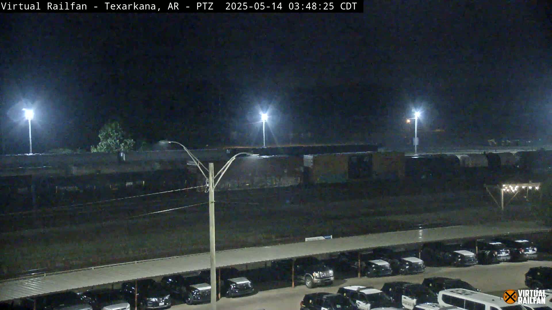 A long freight train sits under bright lights at night next to a parking lot full of cars.
