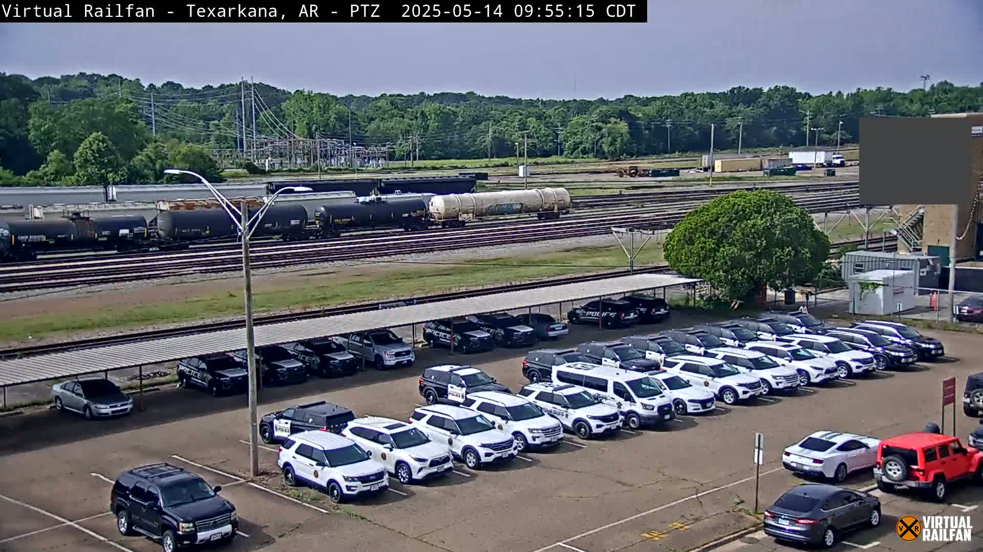 A parking lot filled with numerous police vehicles sits under a covered structure, with a train and a tree-lined horizon visible in the background on a sunny day.