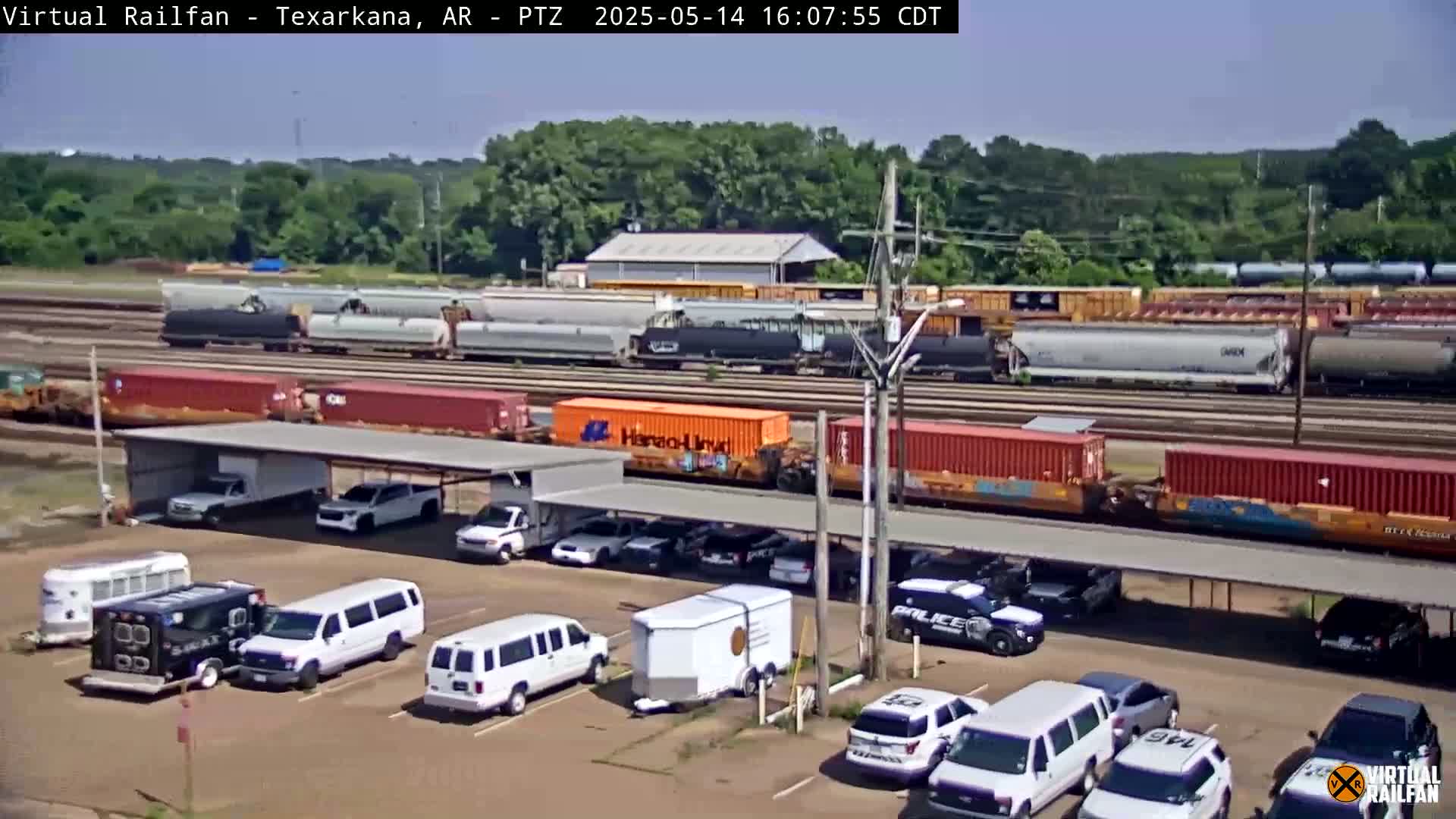 Under a sunny sky, a train carrying various cargo cars passes by a parking lot containing numerous vans, SUVs, and a horse trailer, all situated under a covered structure.
