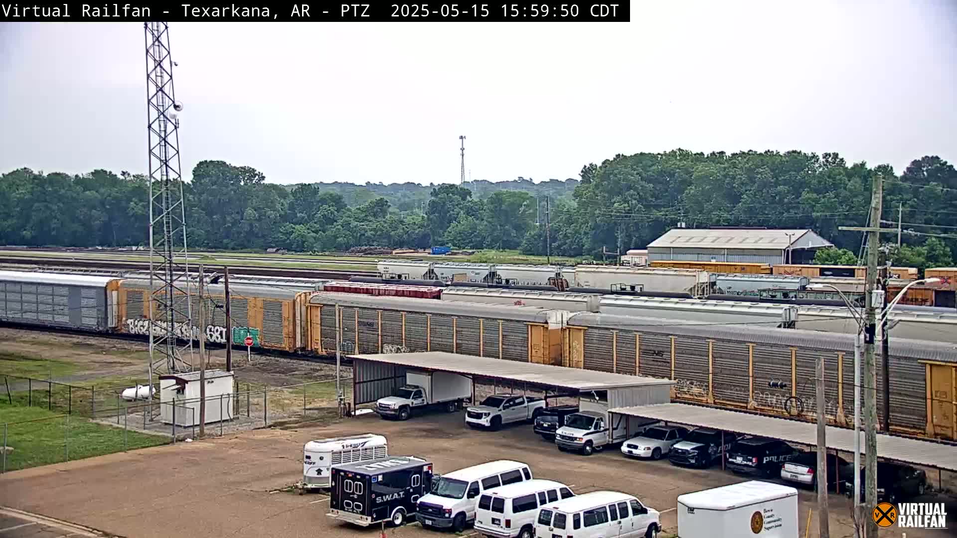 A long freight train is parked near a building with a covered parking lot containing several vehicles, under a partly cloudy sky.
