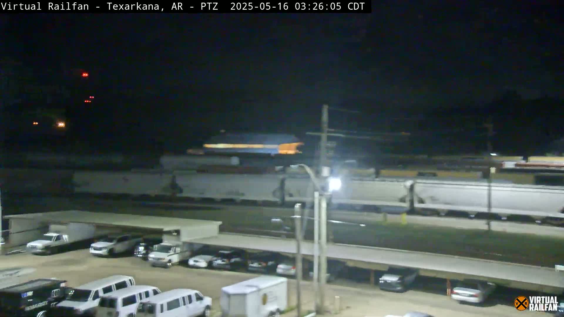 A long freight train passes at night near a parking lot containing several cars and vans.