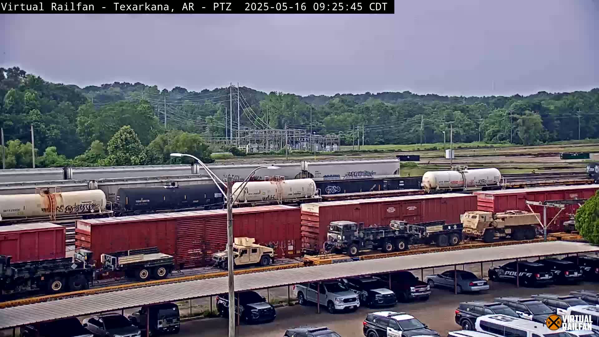 A long freight train carrying military vehicles is parked near a parking lot under an overcast sky.