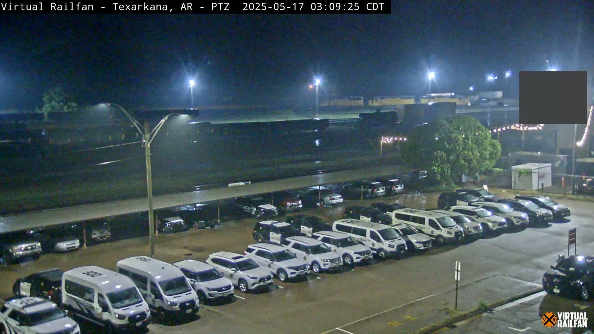 A nighttime view of a wet parking lot filled with mostly white vehicles under covered parking, next to a large dark building under illuminated lights.