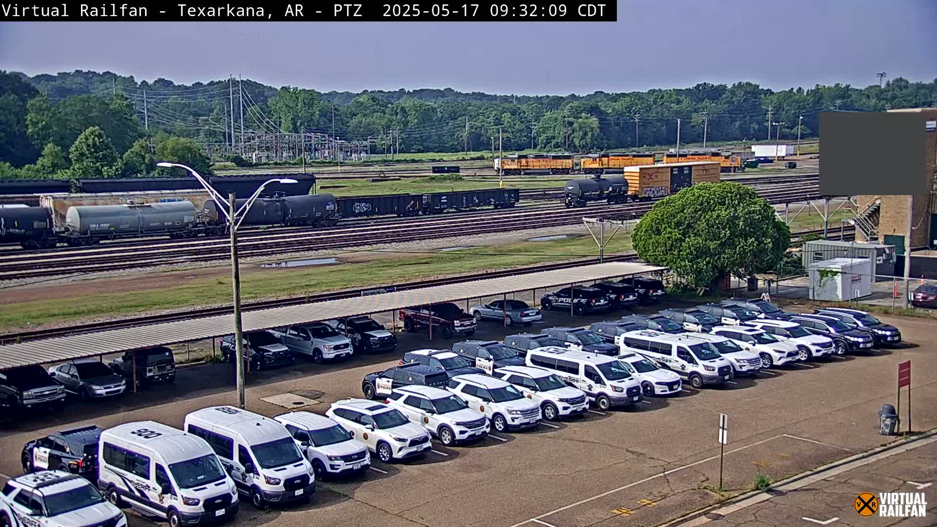 A parking lot filled with a row of white police vehicles is situated near a railroad with several train cars and locomotives under a clear, sunny sky.