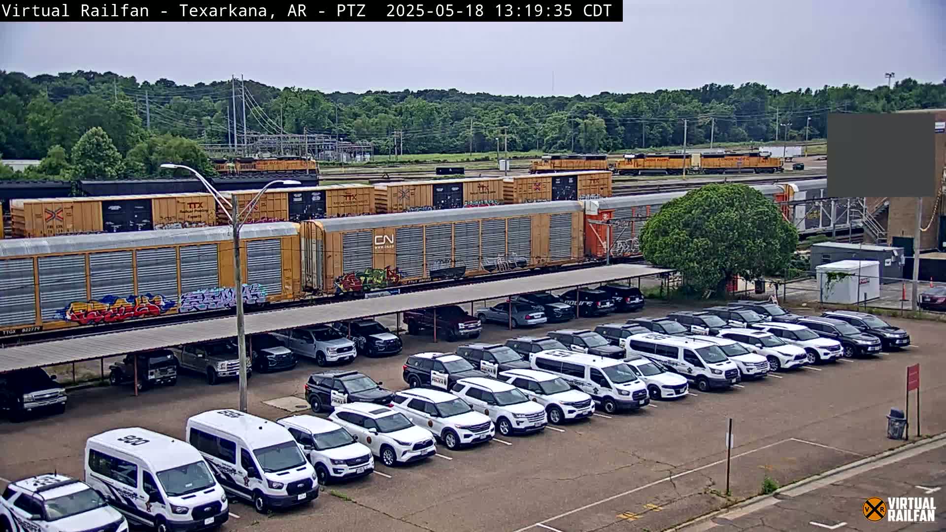 A long freight train sits beside a parking lot filled with mostly white police vehicles under a covered structure, all under an overcast sky.