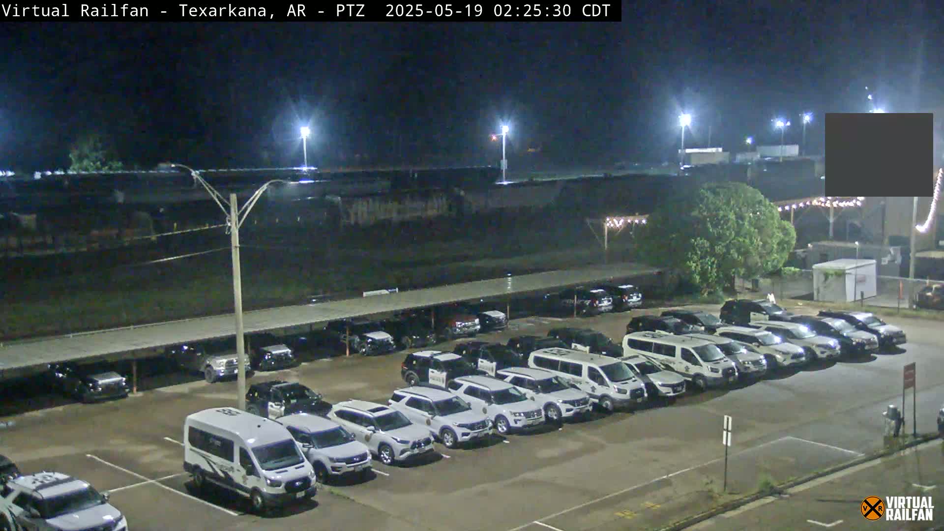 A nighttime view of a large parking lot filled with mostly white vehicles under a covered structure, next to a building and under lights.