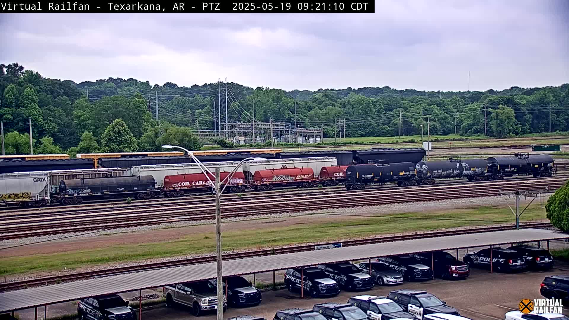 A long freight train sits on railroad tracks under an overcast sky, with a parking lot full of police vehicles in the foreground.