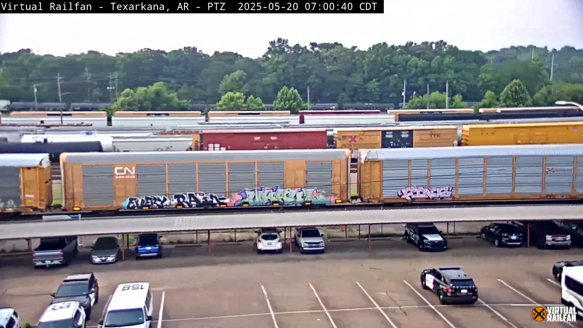 A freight train with graffiti on some of its cars passes by a parking lot with several cars and police vehicles under a covered structure on a partly sunny day.