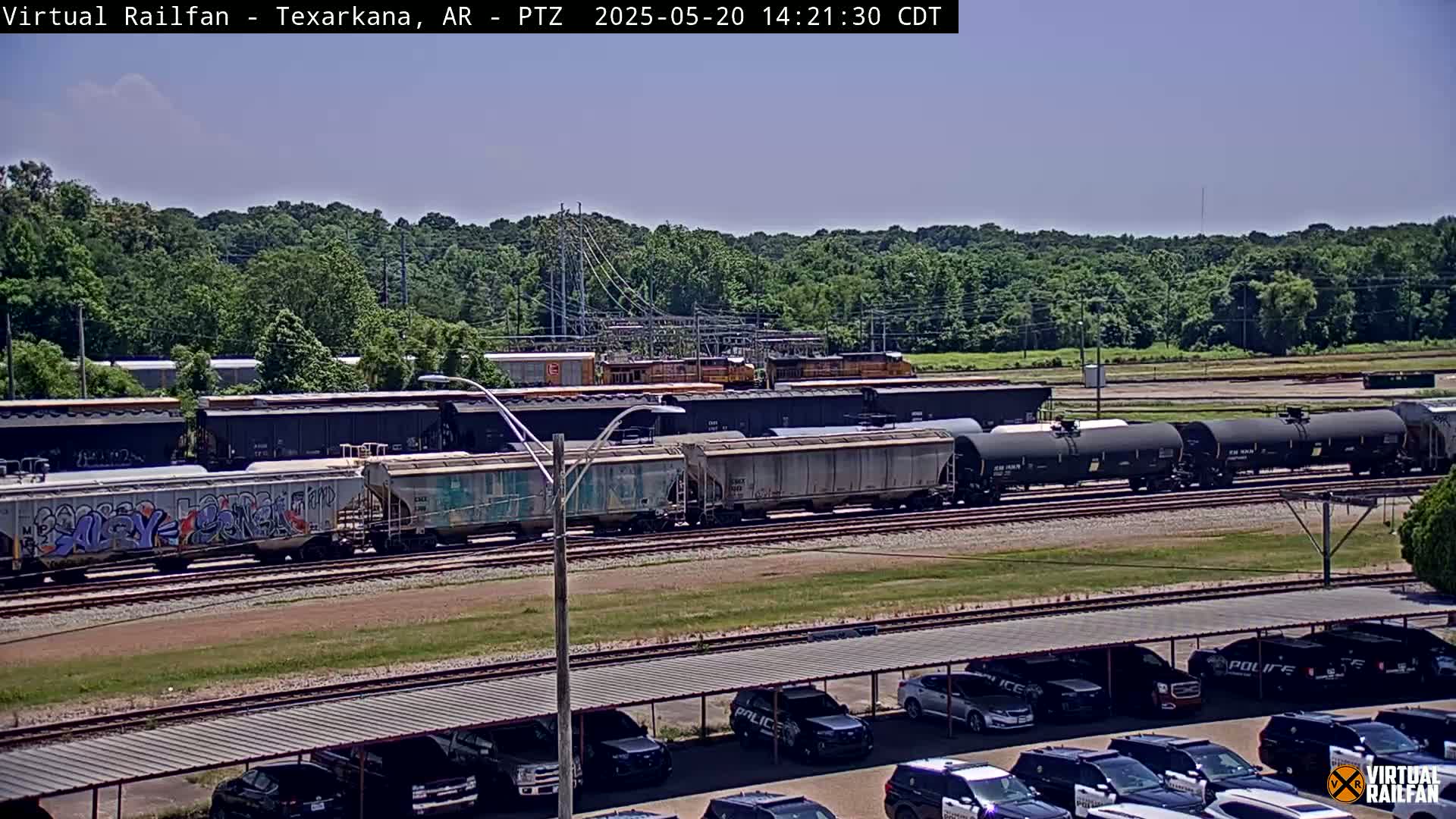 A long freight train sits on railroad tracks under a clear, sunny sky, with a parking lot full of police vehicles in the foreground.