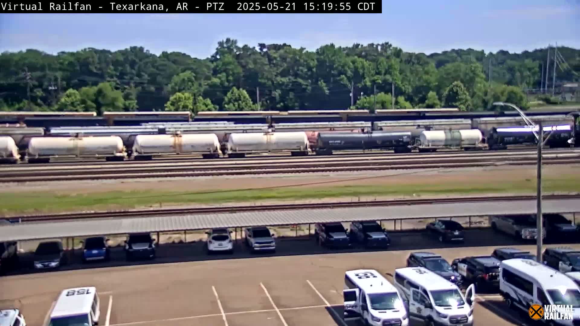 A long freight train carrying tankers is parked on railroad tracks behind a parking lot with several cars and vans under a covered shelter, on a sunny day.