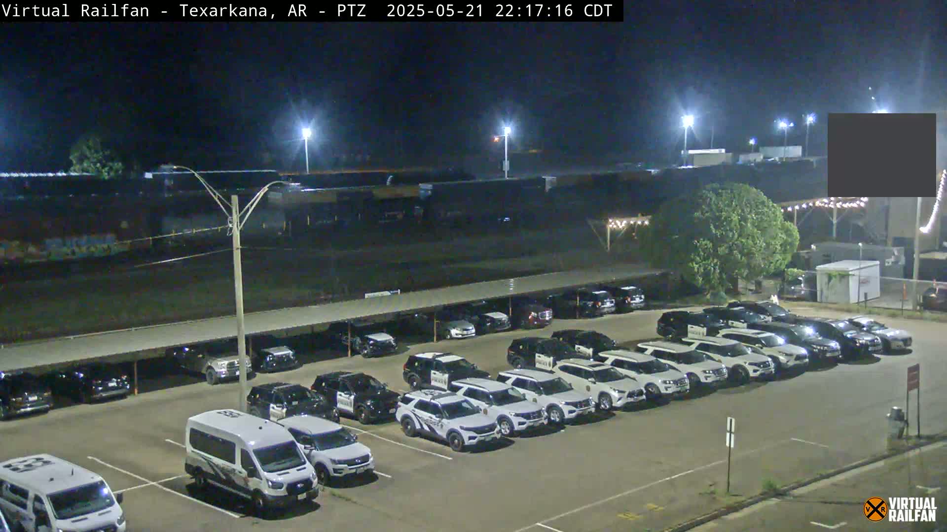 A nighttime view of a parking lot filled with mostly white SUVs, under a covered structure, adjacent to a rail yard.
