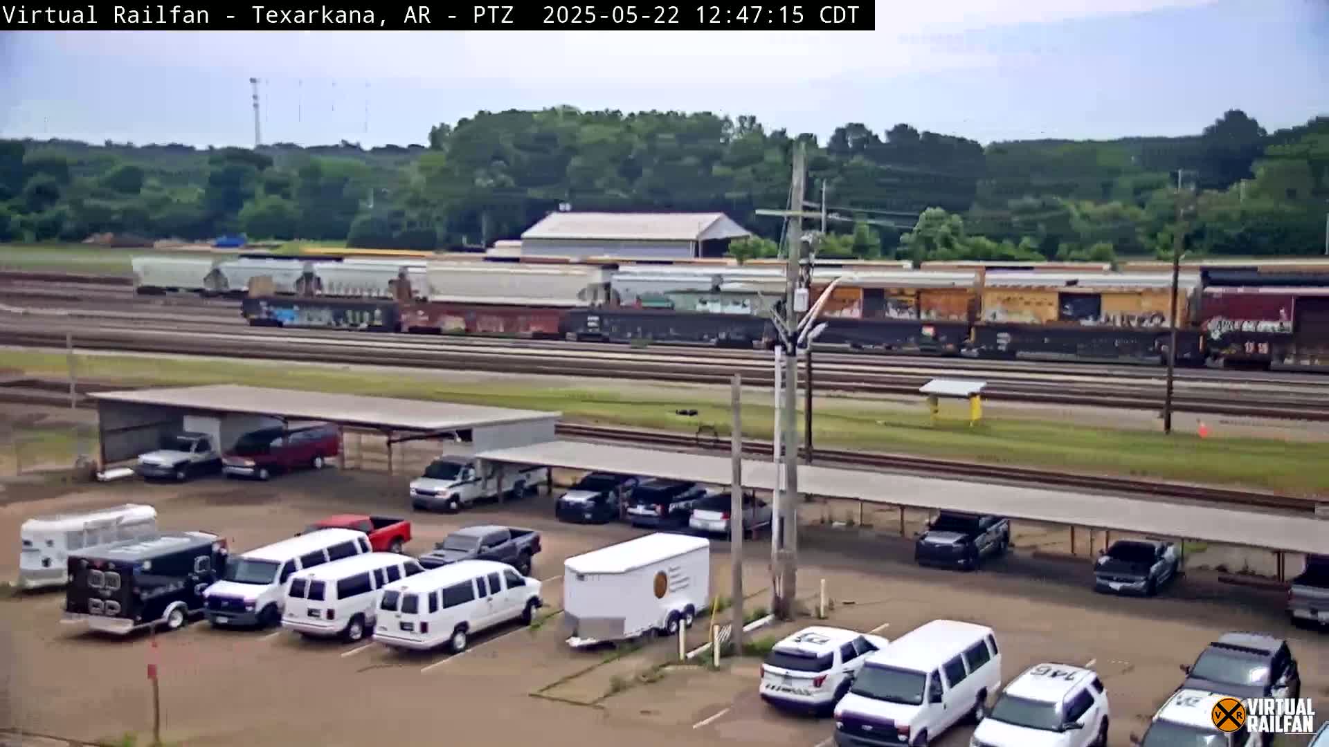 A long freight train passes by a parking lot with several vans and cars under a covered structure on a partly sunny day.