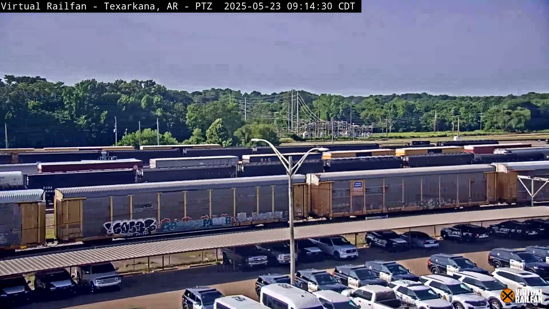 A long line of freight cars sits in a rail yard on a sunny day, next to a parking lot filled with police vehicles and other cars under a covered structure.
