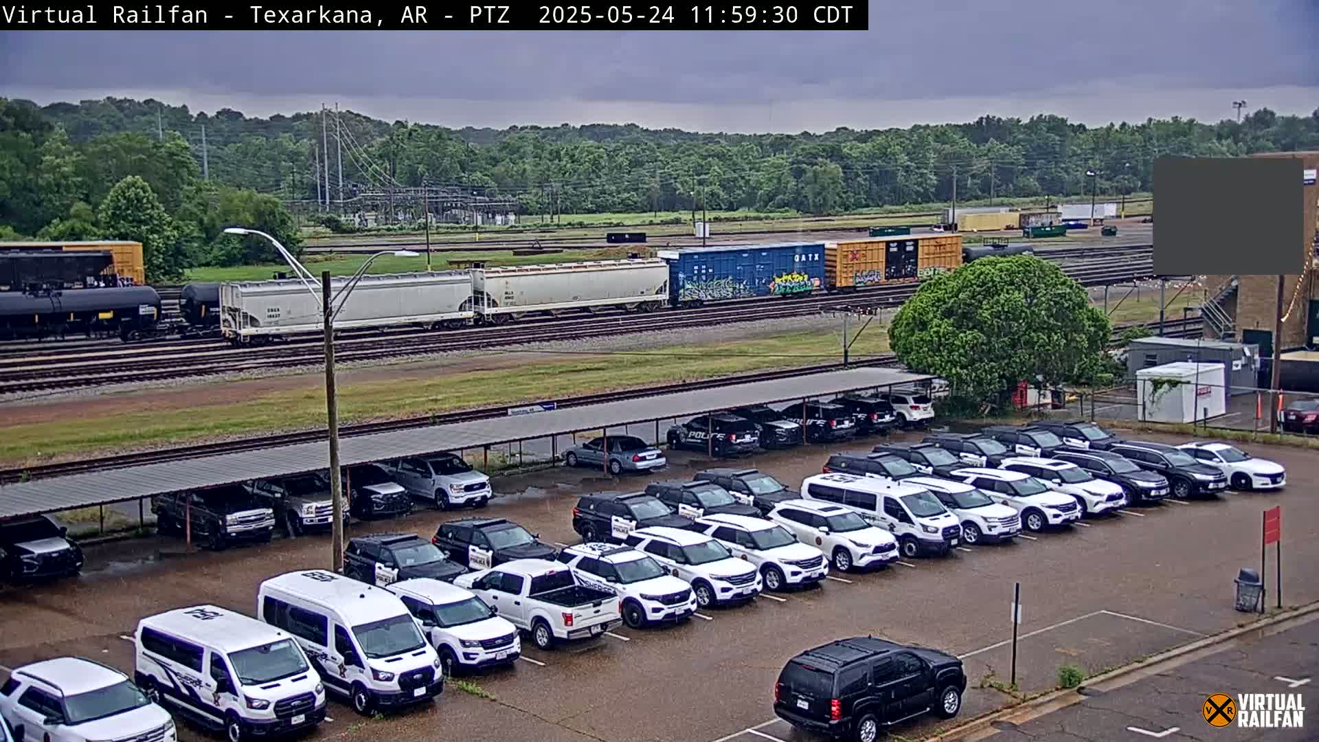 Under an overcast sky, a train passes by a parking lot filled with mostly white police vehicles parked under a covered structure.