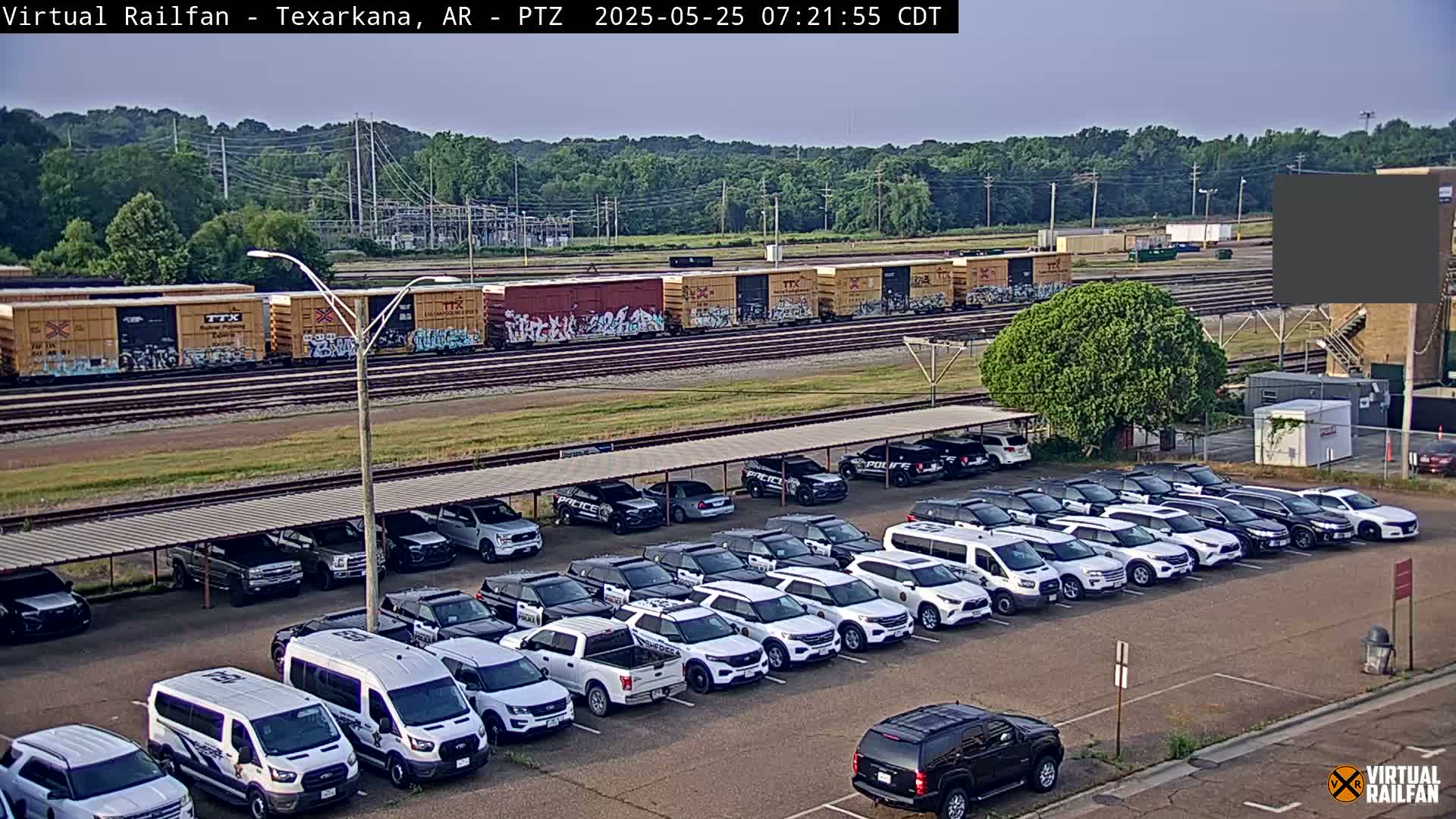 A long train of graffitied boxcars sits on railroad tracks behind a large parking lot full of mostly white police vehicles under a covered structure, all under a mostly clear sky.