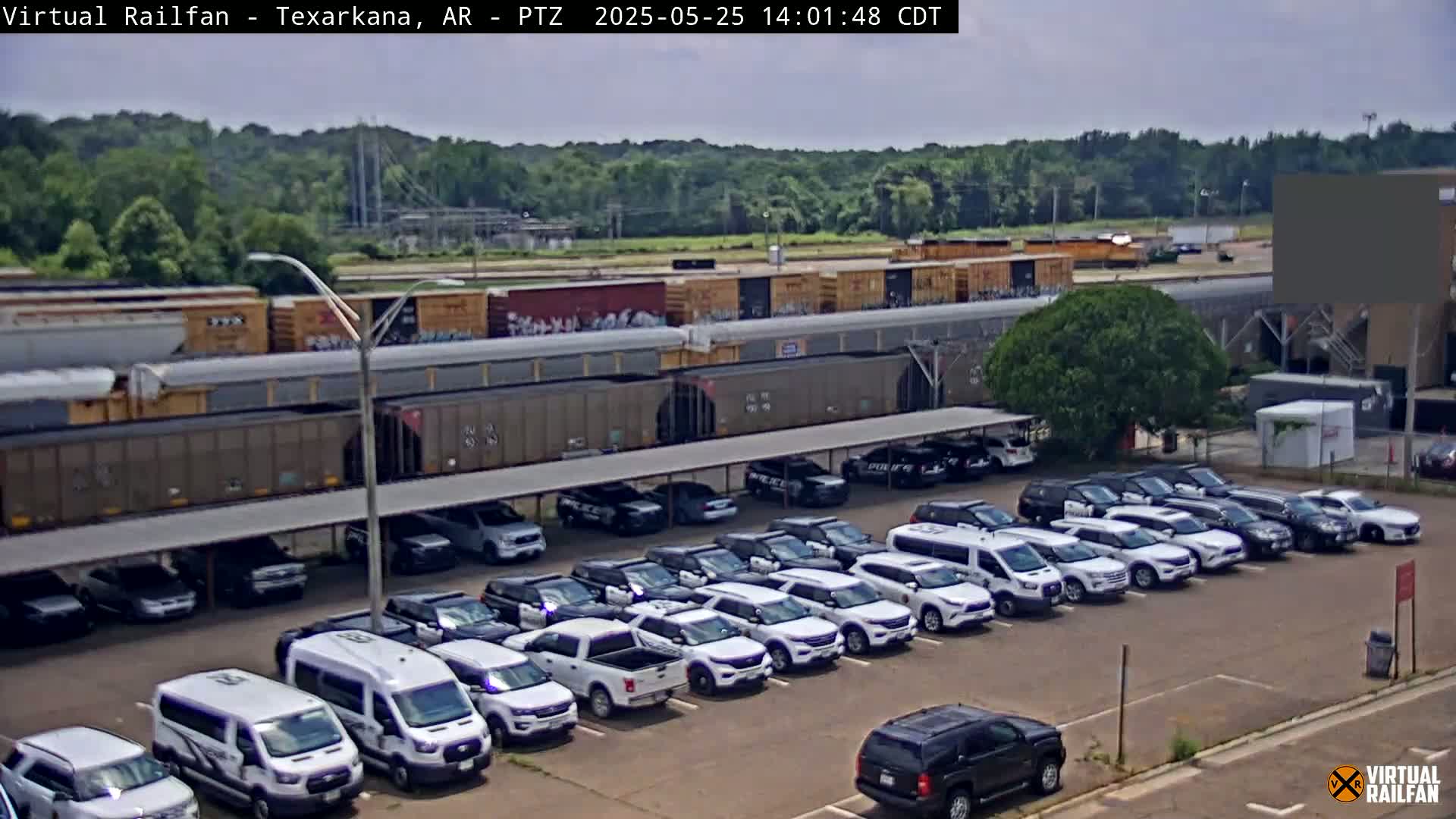 A long freight train sits behind a parking lot full of mostly white SUVs and vans under a covered structure, on a partly cloudy day.