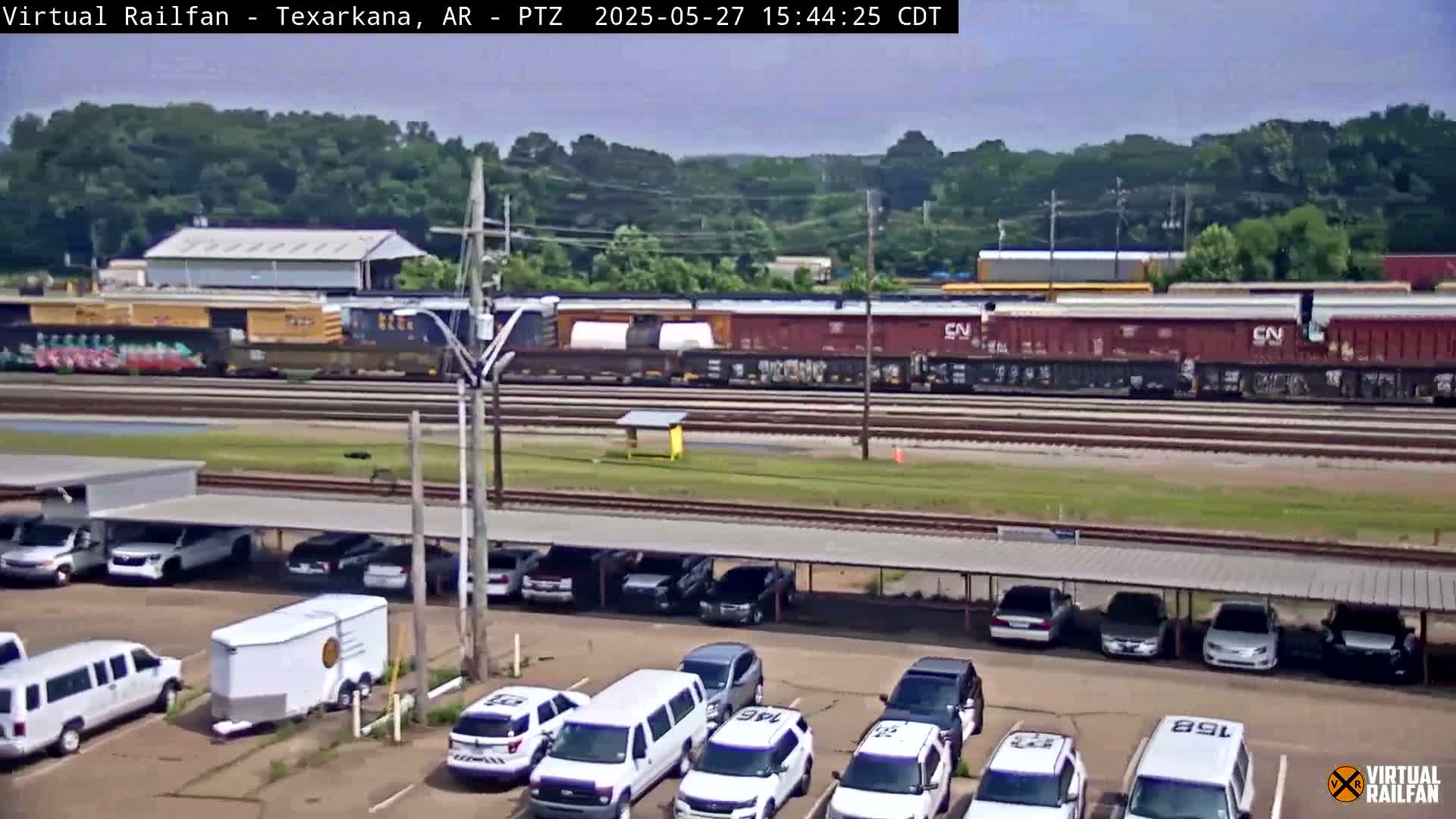 A long freight train passes by a parking lot filled with cars and vans under a covered structure on a sunny day.