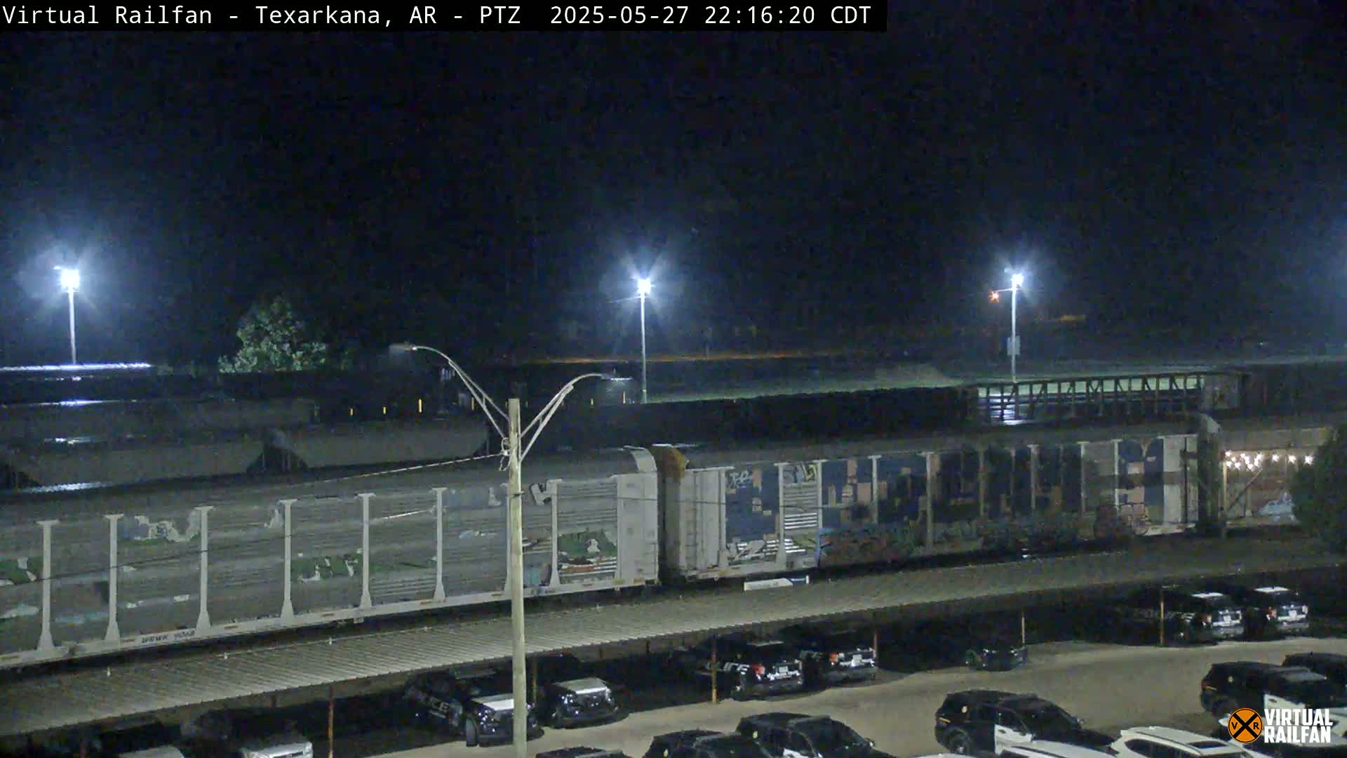 At night, a freight train with graffiti on some cars sits in a rail yard next to a parking lot with numerous vehicles under a covered structure.