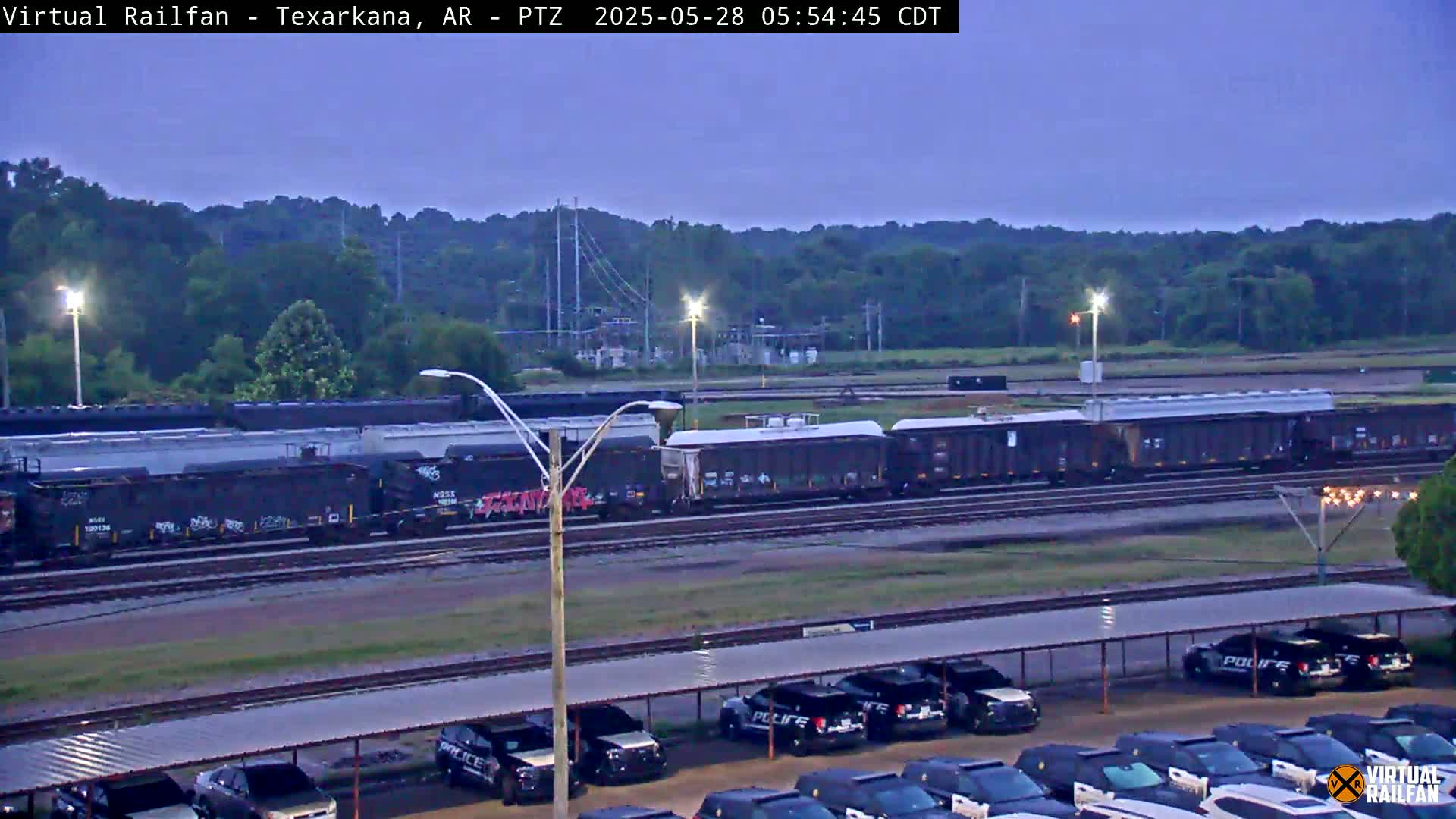 A long freight train sits on railroad tracks under a twilight sky, with a parking lot full of police vehicles visible in the foreground.