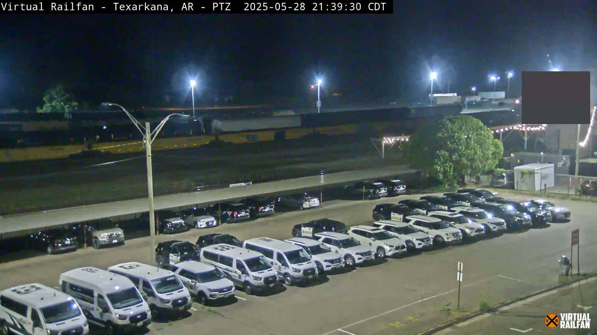 A nighttime view of a parking lot filled with mostly white SUVs and vans under a covered structure, with a long freight train visible in the background under partly cloudy skies.