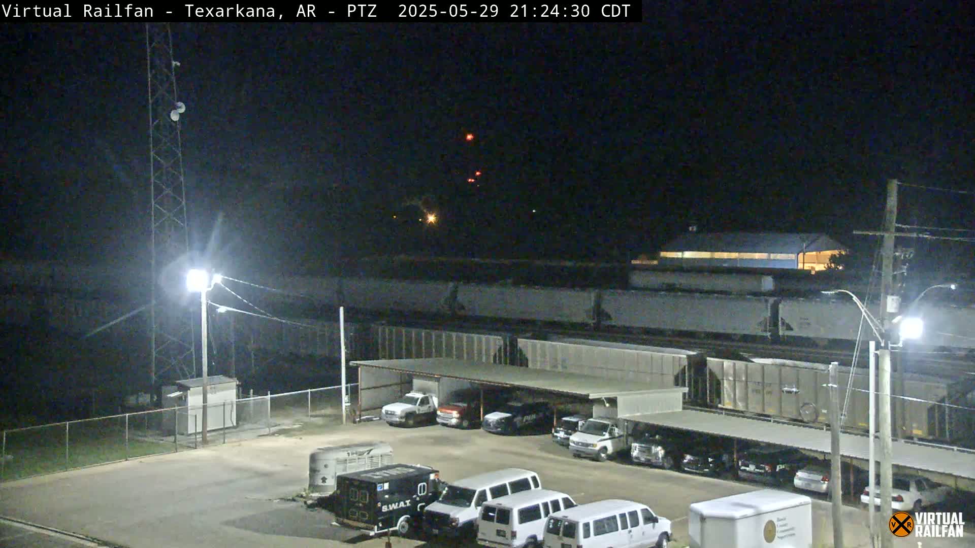 At night, a long freight train is parked near a parking lot containing several vehicles under a covered structure.
