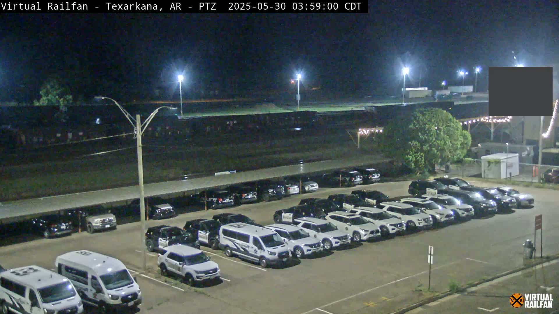 A nighttime view of a large parking lot filled with mostly white SUVs and vans under a covered structure, adjacent to a field and some buildings under dark, clear skies.