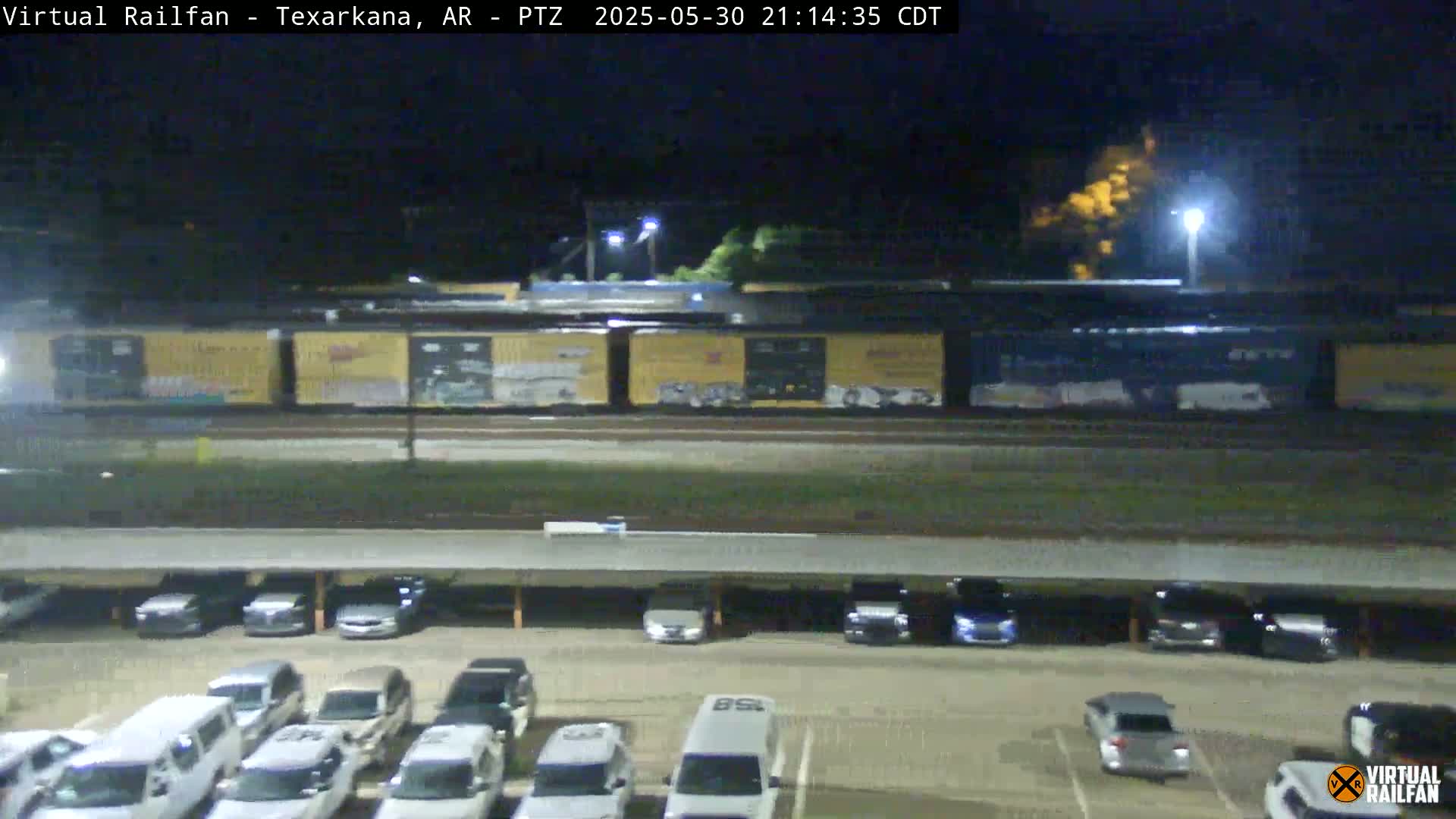 A long freight train passes by a mostly empty parking lot at night.