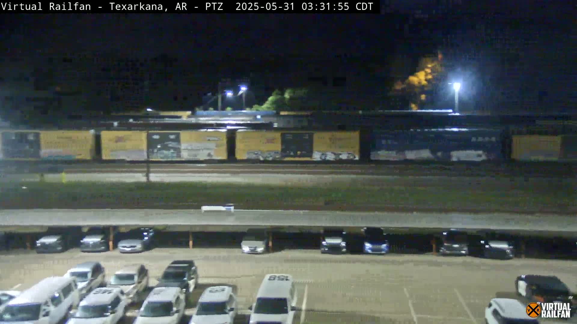 At night, a long freight train passes by a mostly empty parking lot under a covered structure.