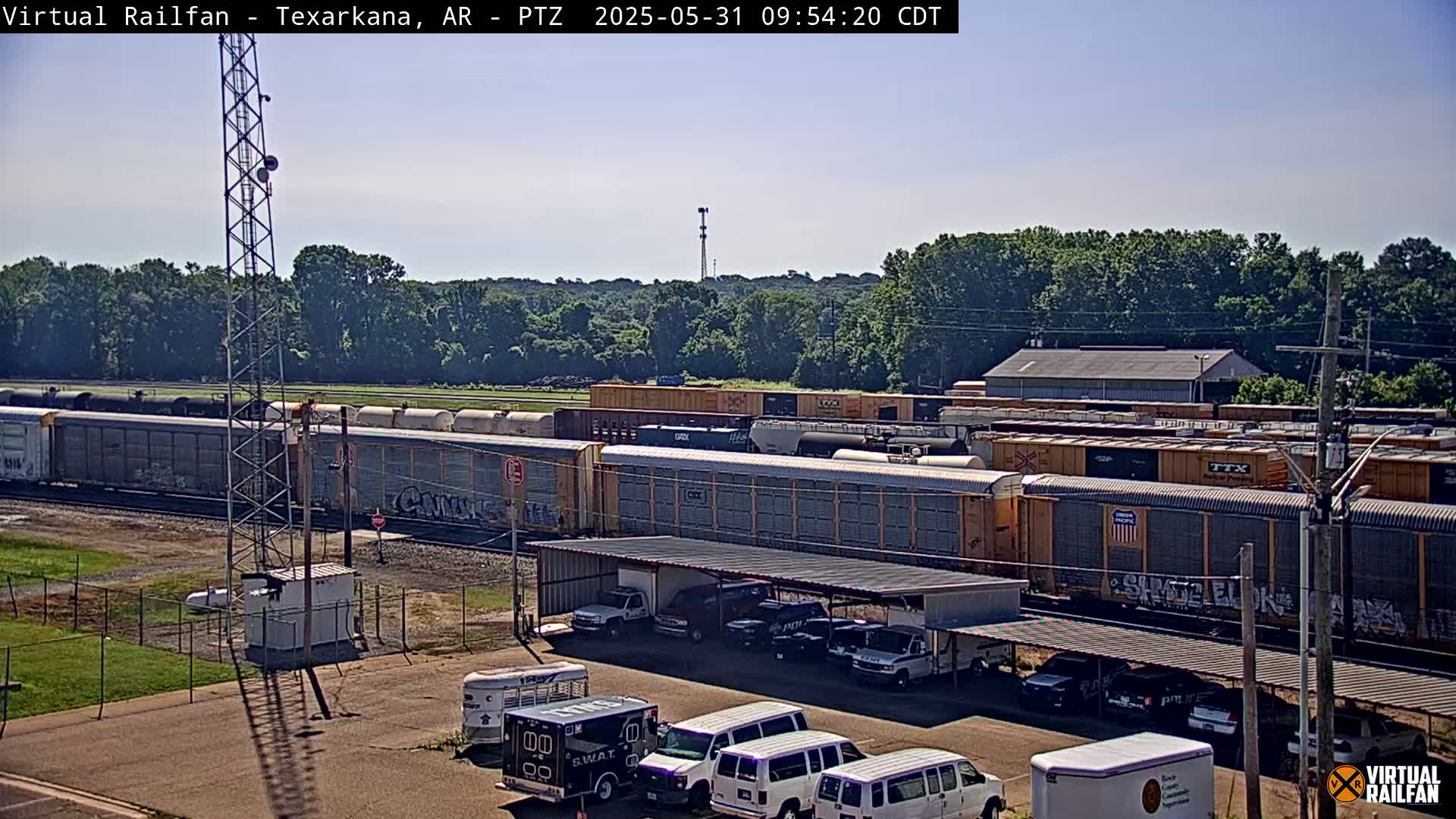 A long freight train sits on railroad tracks under a clear, sunny sky, next to a parking lot containing several vehicles, including a SWAT van.