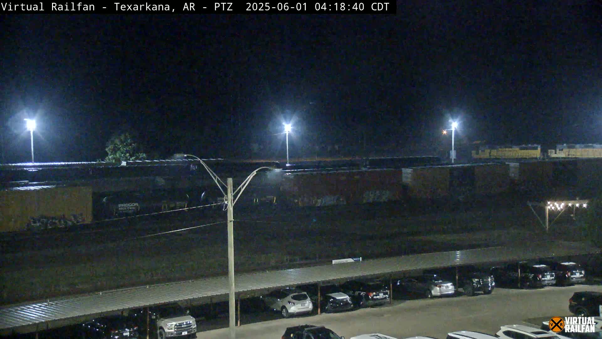 A nighttime view of a train car, graffiti-covered rail cars, and a parking lot with cars under a covered structure.