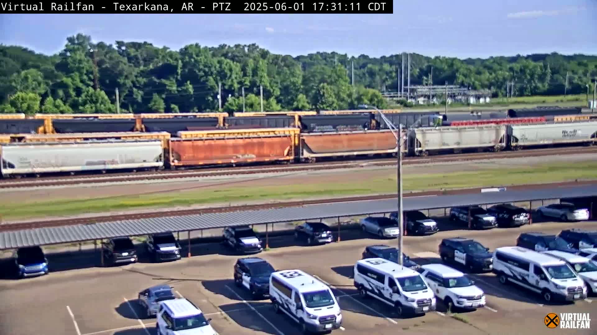 A long freight train passes by a parking lot full of mostly white vans under a covered structure on a sunny day.