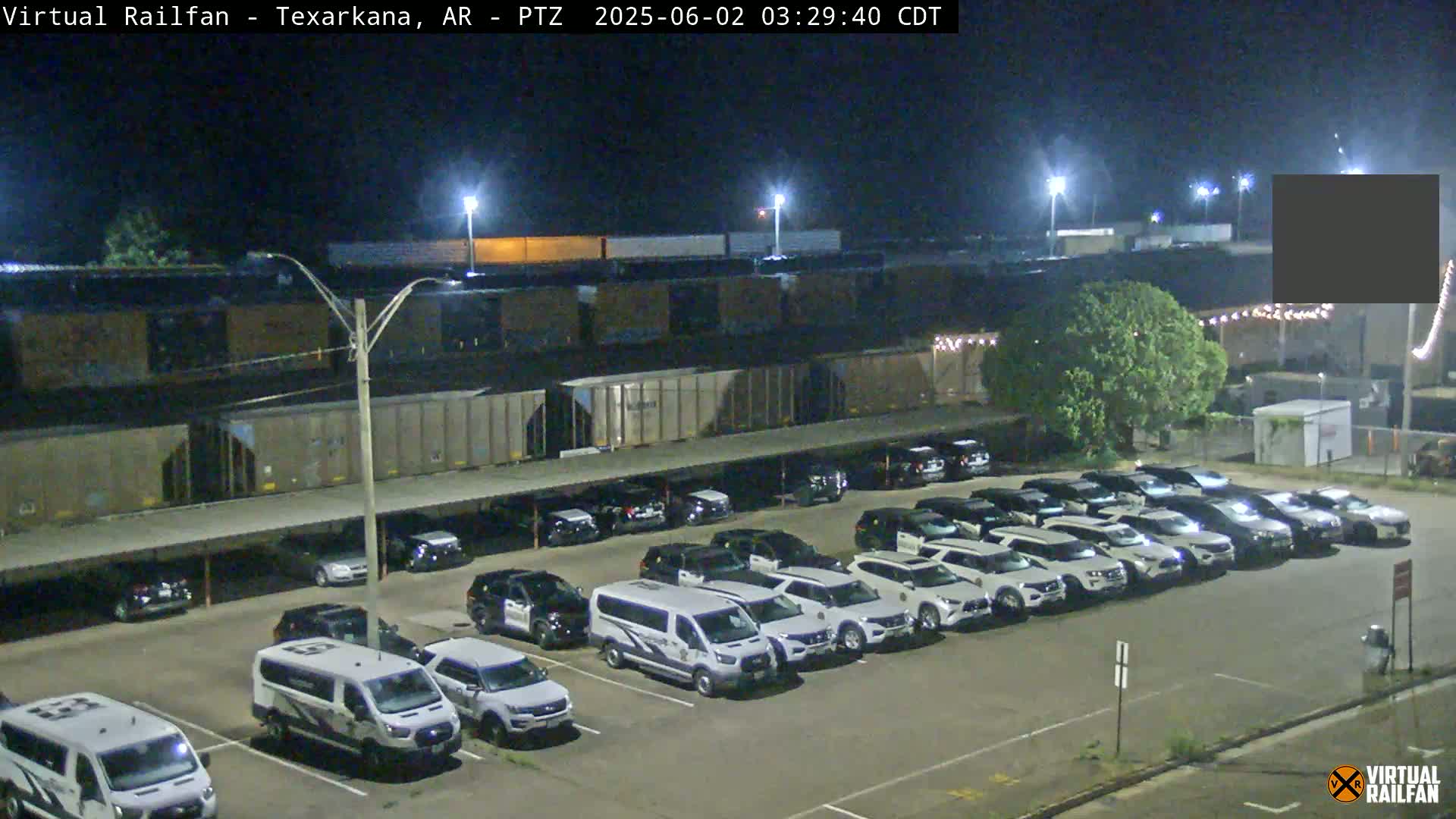 A nighttime view of a large parking lot filled with mostly white SUVs and vans, situated next to a train and some industrial buildings under bright lights.