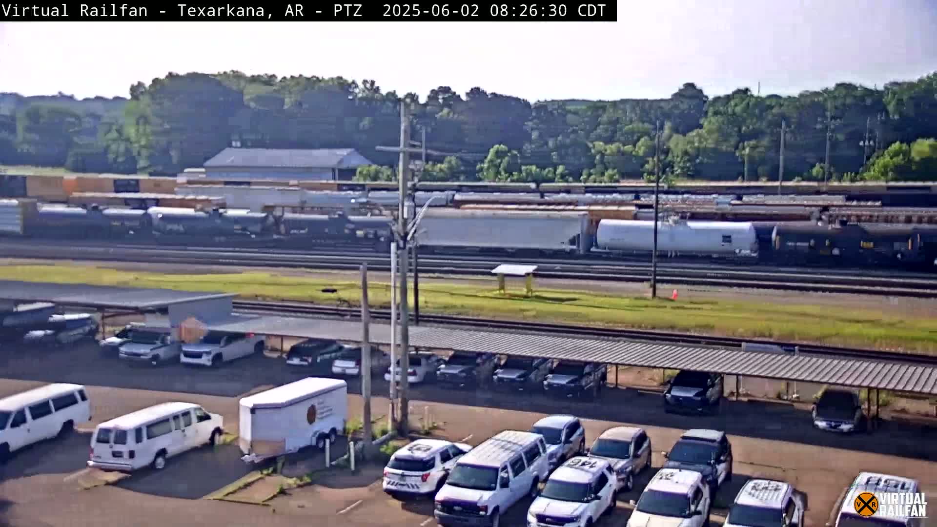 A long freight train passes by a parking lot filled with cars and vans under a covered structure on a sunny day.