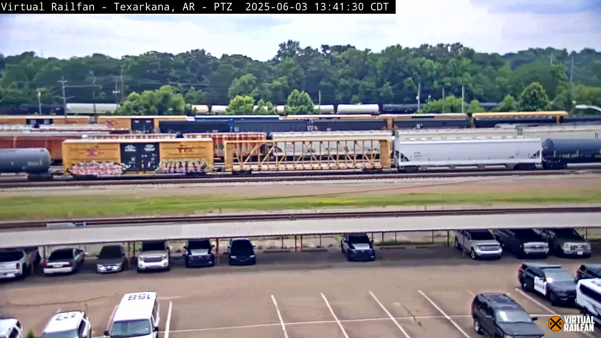 A long freight train sits on tracks in front of a parking lot with several cars parked under a covered structure, all under a partly cloudy sky.
