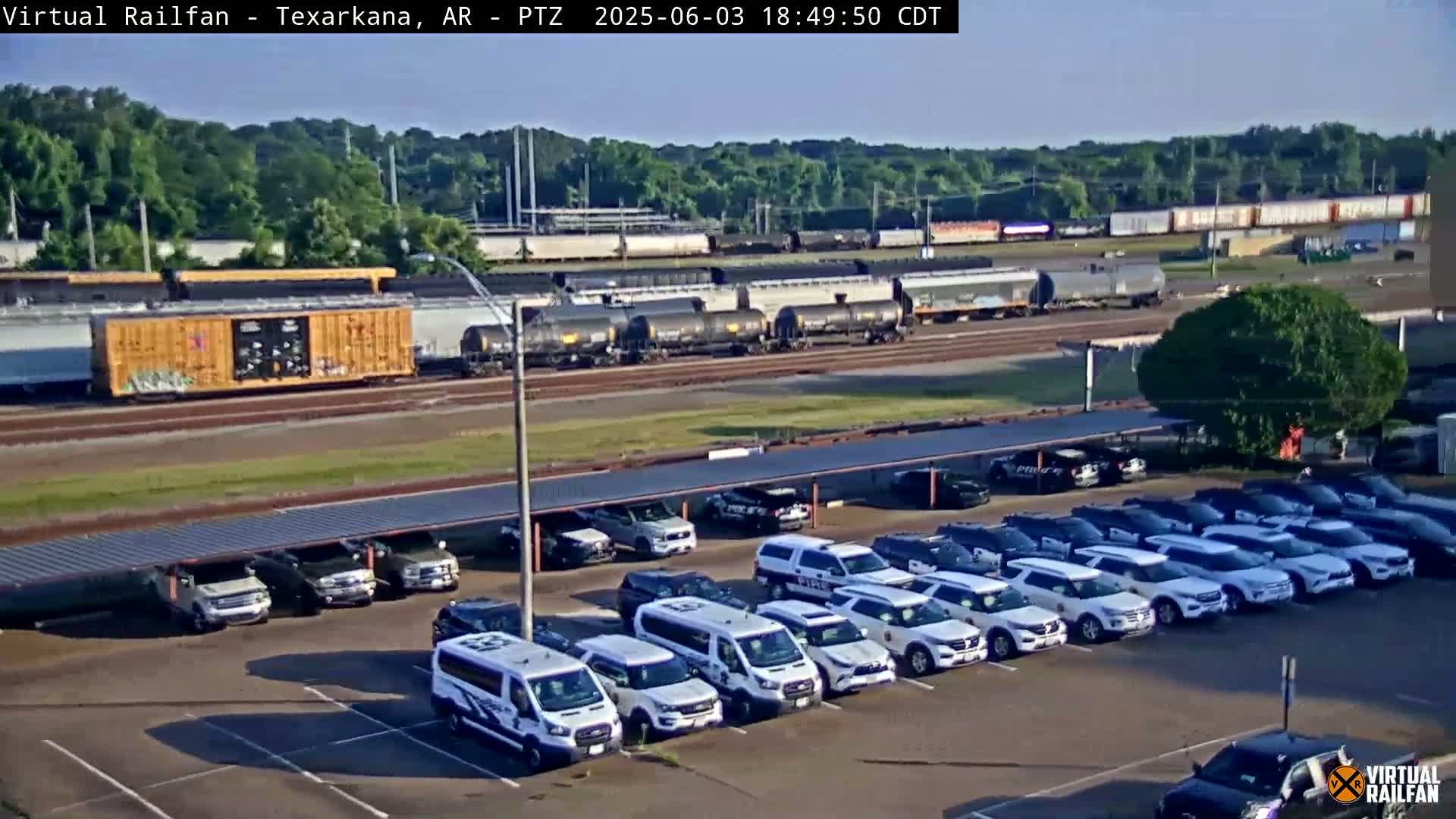 A long freight train is visible in the distance behind a large parking lot filled with numerous white SUVs under a covered structure, all on a partly sunny day.